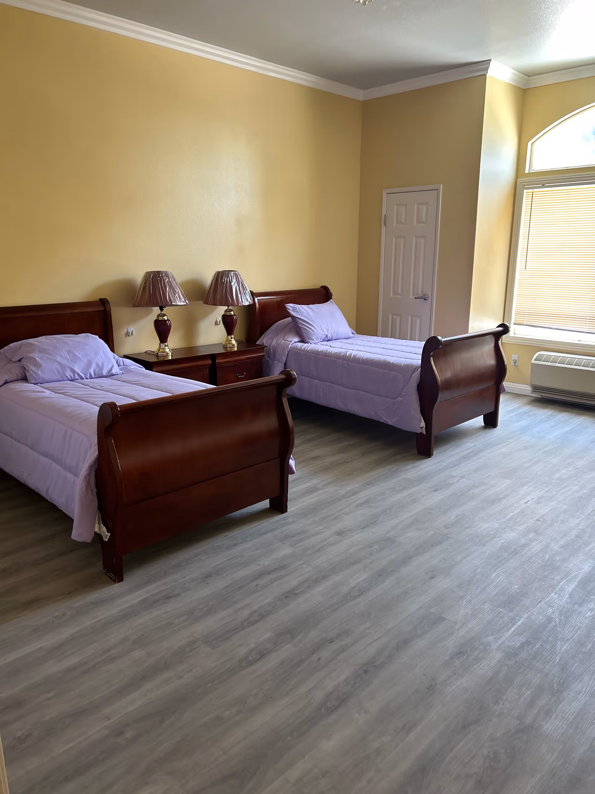 A bedroom with two wooden twin beds, each made with light purple bedding and pillows. Between the beds is a wooden nightstand with two matching lamps that have dark red bases and are covered with plastic. The room has light yellow walls, a large window with blinds, and light gray wood flooring.