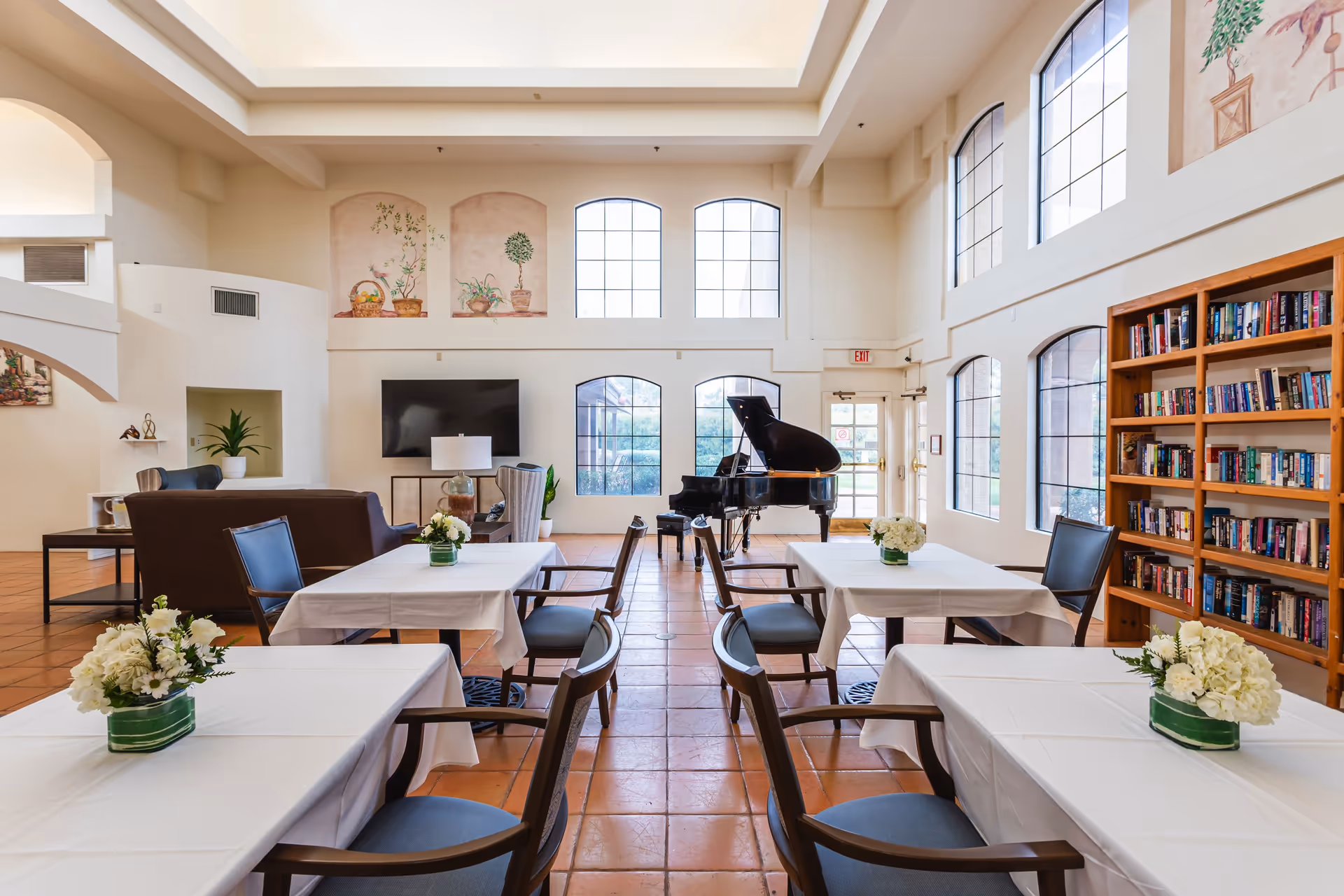 Bright communal dining and lounge area with tables set with white tablecloths and floral centerpieces, a grand piano, bookshelves, and large arched windows.