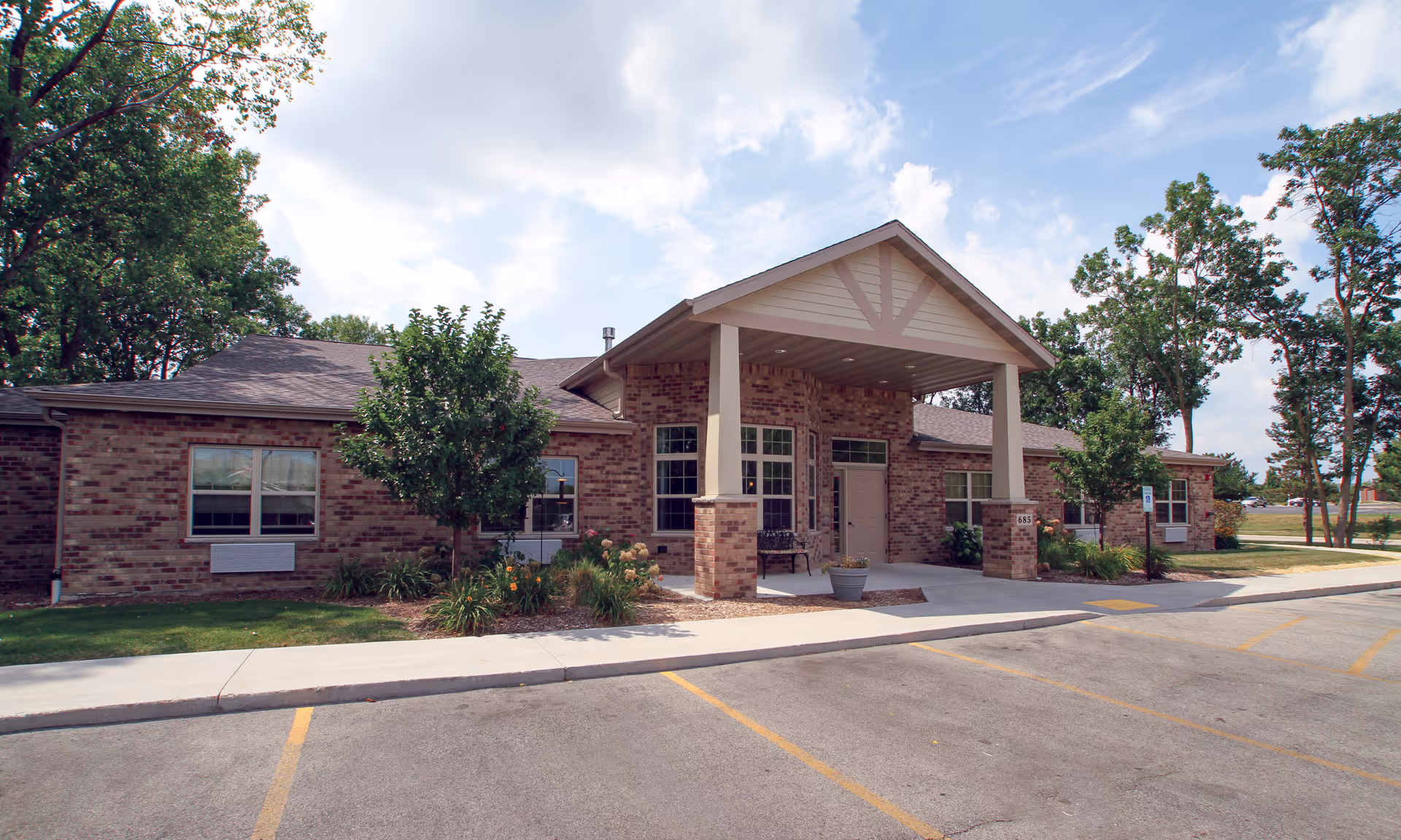 Exterior view of a single-story brick building with a covered entrance supported by two columns. There are trees and landscaping around the building, a sidewalk leading to the entrance, and a parking lot with marked spaces in front. The sky is partly cloudy.