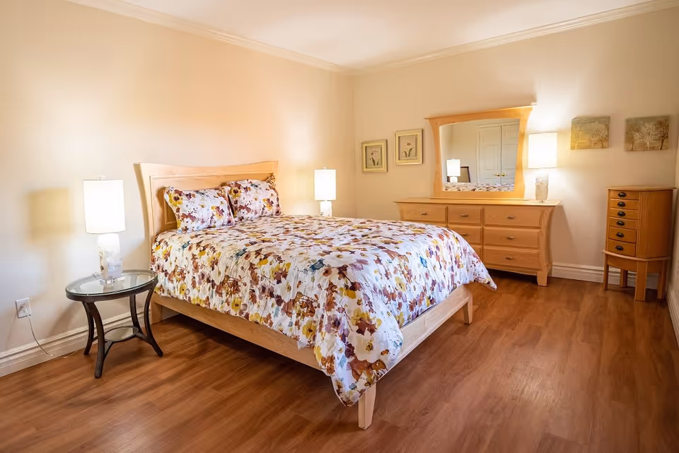 A bright bedroom with a wooden bed dressed in floral bedding, two lamps, a dresser with mirror, and hardwood floors.