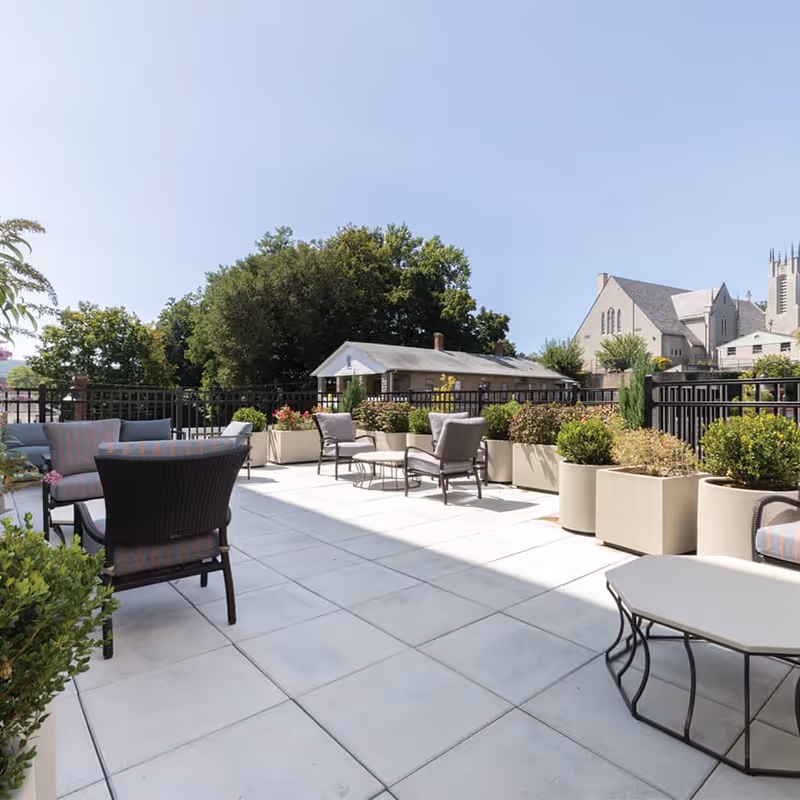 Outdoor patio area with cushioned chairs and small tables arranged on a tiled floor, surrounded by large planters with green shrubs and plants, under a clear blue sky with trees and buildings in the background.