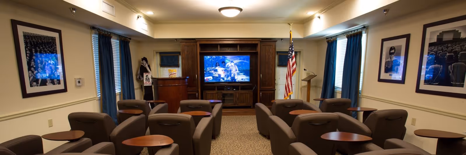 A small theater-style room with multiple rows of brown armchairs, each with a small attached wooden table. At the front of the room is a large flat-screen TV mounted on a wooden cabinet, an American flag, a podium, and a popcorn machine. The walls have framed black and white photos, and windows with blue curtains.