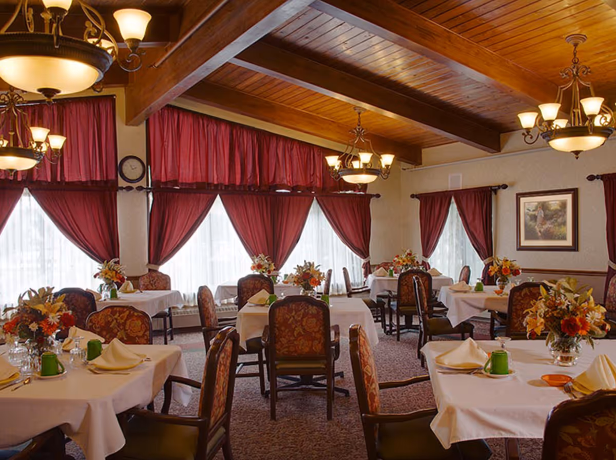 A dining room with multiple tables covered in white tablecloths, each set with folded napkins, green cups, and floral centerpieces. The room features wooden ceiling beams, ornate chandeliers, and large windows with red curtains allowing natural light to filter in. Upholstered chairs with patterned fabric surround the tables, and a framed painting hangs on one wall.