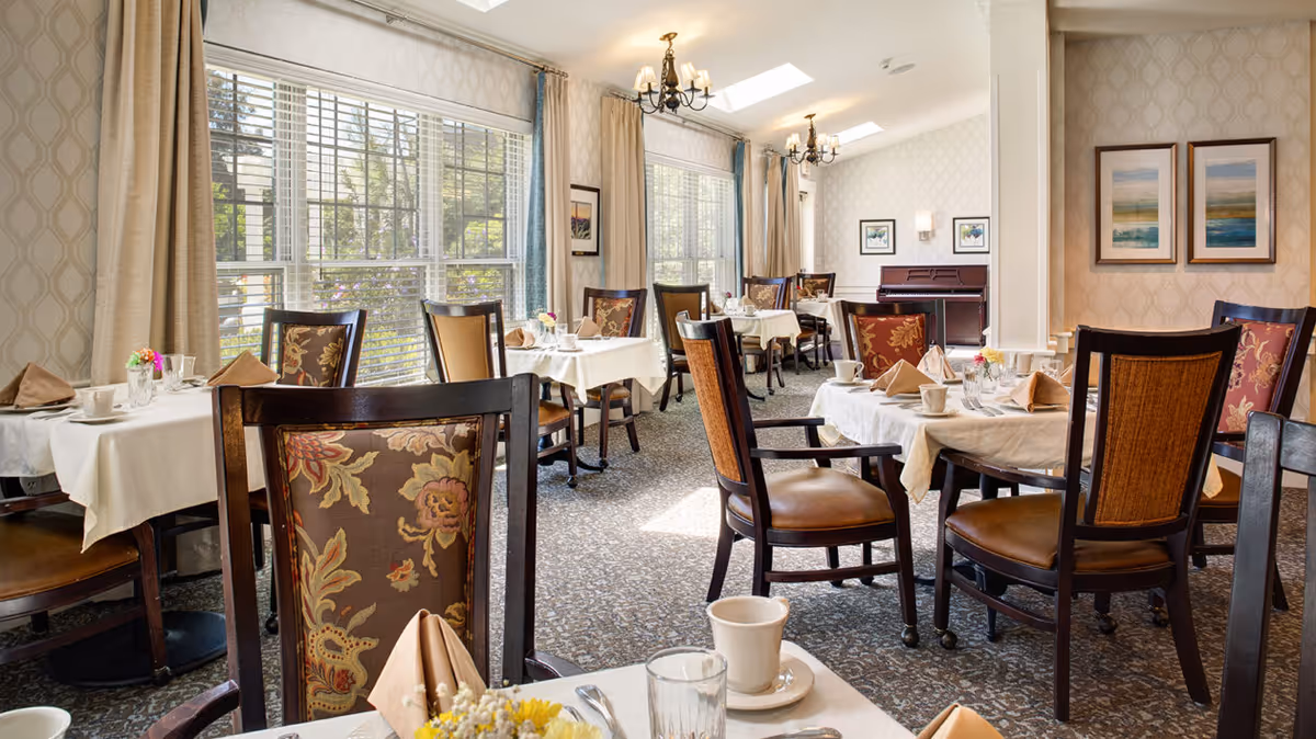A bright and elegant dining room with multiple tables covered in white tablecloths, each set with cups, glasses, napkins, and small flower arrangements. The room features large windows with beige curtains allowing natural light to fill the space. The chairs have dark wooden frames with floral and patterned upholstery. The walls are decorated with framed artwork, and there are two chandeliers hanging from the ceiling.