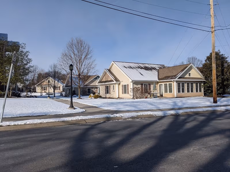 A residential-style building with beige siding and a snow-covered roof, surrounded by a snowy lawn and leafless trees under a clear blue sky. A sidewalk and street are visible in the foreground.