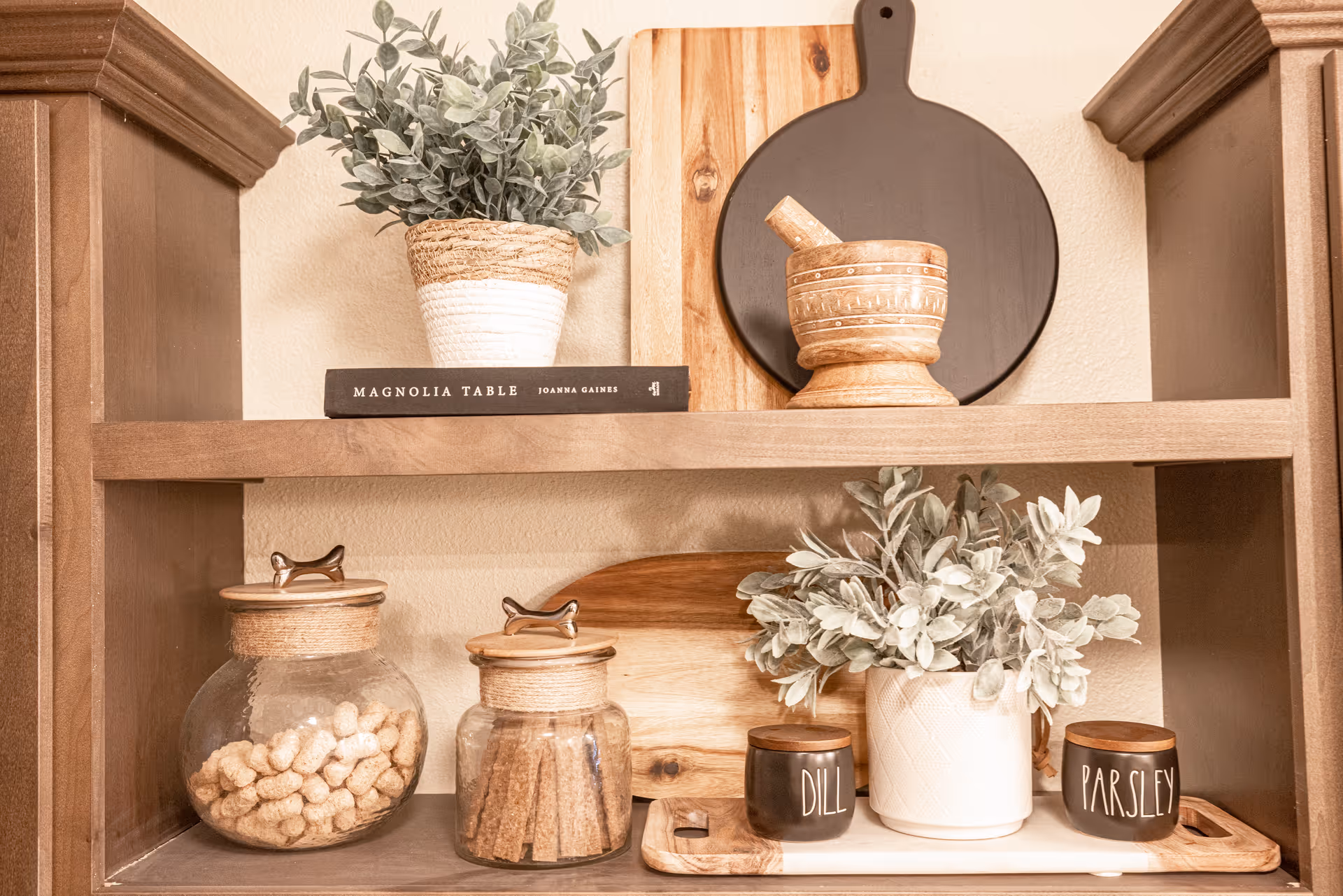 Wooden shelves with decorative items including a potted plant, a book titled Magnolia Table by Joanna Gaines, a wooden mortar and pestle, two glass jars with wooden lids containing snacks, a wooden cutting board, a white vase with greenery, and two small containers labeled Dill and Parsley.