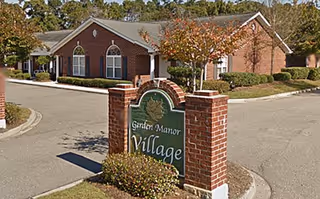 Exterior view of a brick building with a peaked roof and arched windows, surrounded by trees and landscaping. In the foreground, there is a brick signpost with a green sign that reads 'Garden Manor Village.'