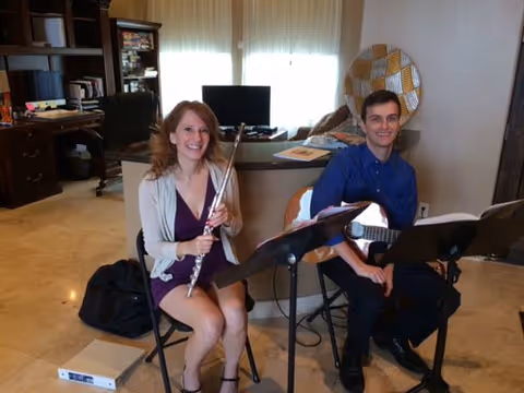 Two people sitting indoors playing musical instruments. A woman on the left is holding a flute, and a man on the right is holding a guitar. They are seated on chairs with music stands in front of them. The room has a desk, a computer monitor, and a decorative wall piece in the background.