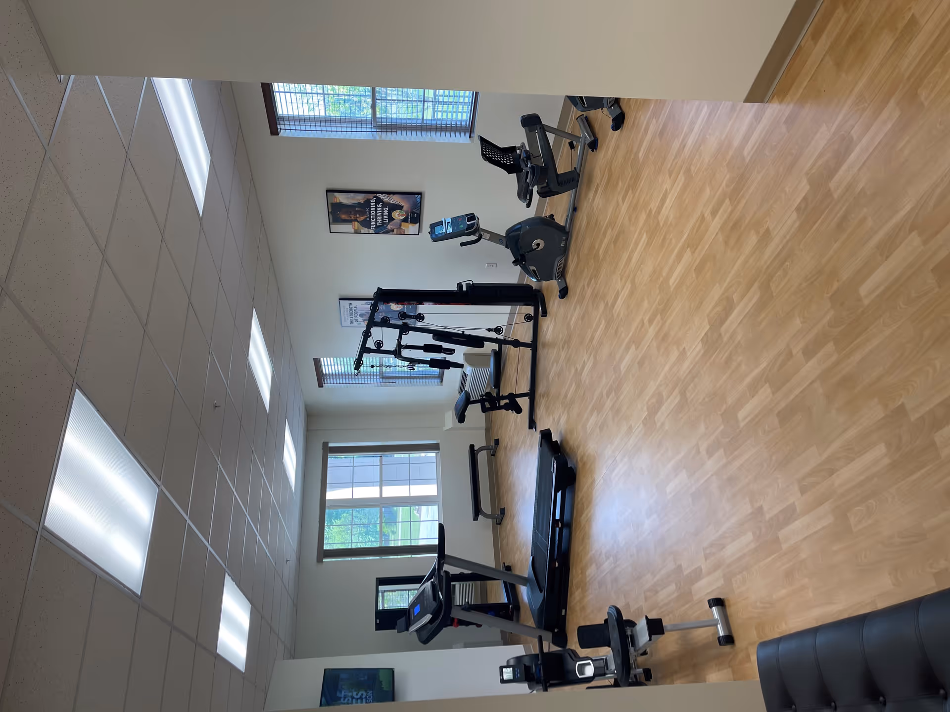 A fitness room with exercise equipment including a treadmill, stationary bike, and multi-function weight machine. The room has wooden flooring, large windows with blinds, and motivational posters on the walls.
