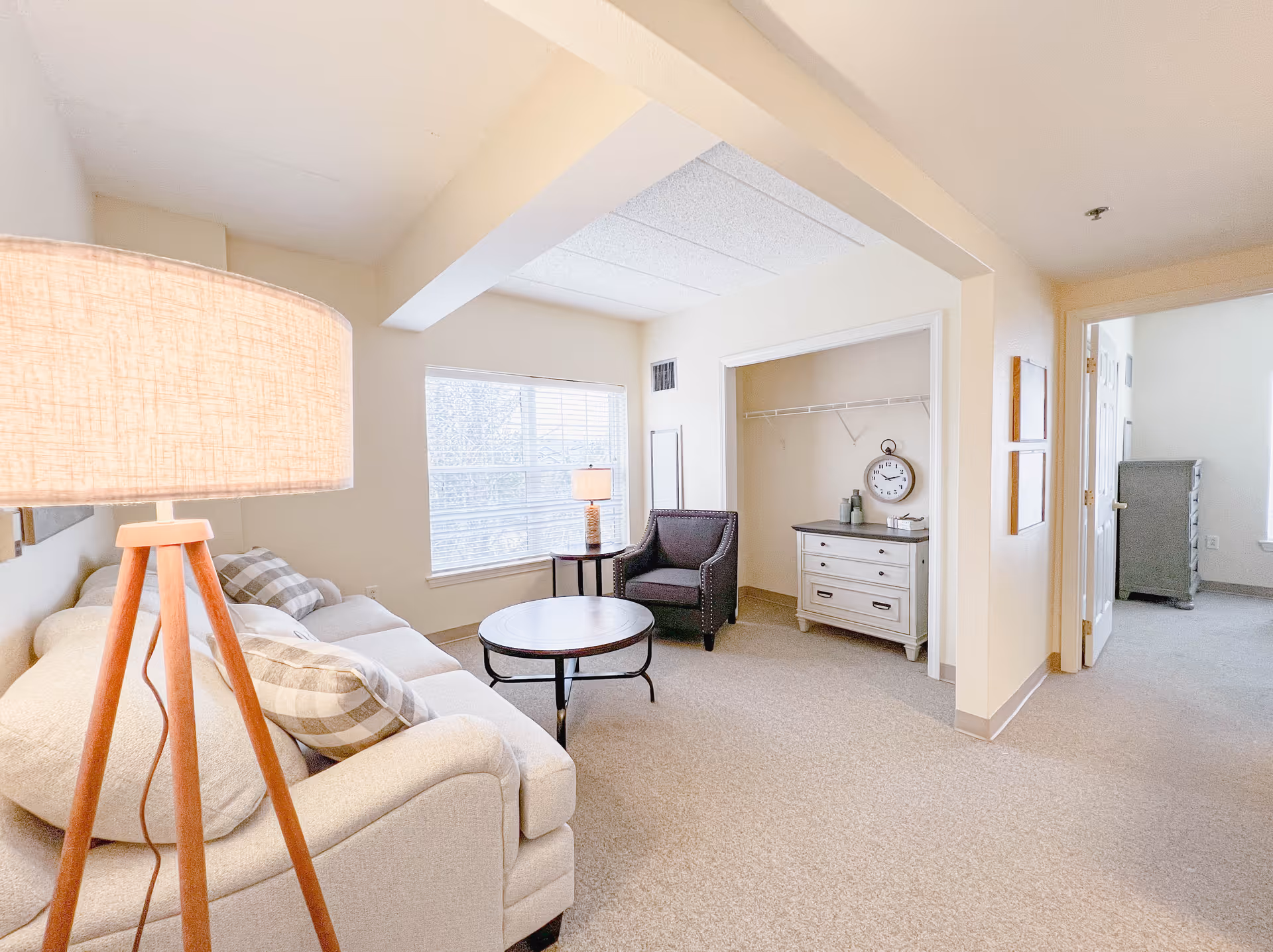 A bright and cozy living room area in a senior living facility featuring a beige sofa with plaid pillows, a round wooden coffee table, a dark armchair, and a floor lamp with a wooden tripod base. There is a large window letting in natural light, and an open closet space with a white dresser and a wall clock. The room has light-colored walls and carpeted flooring.