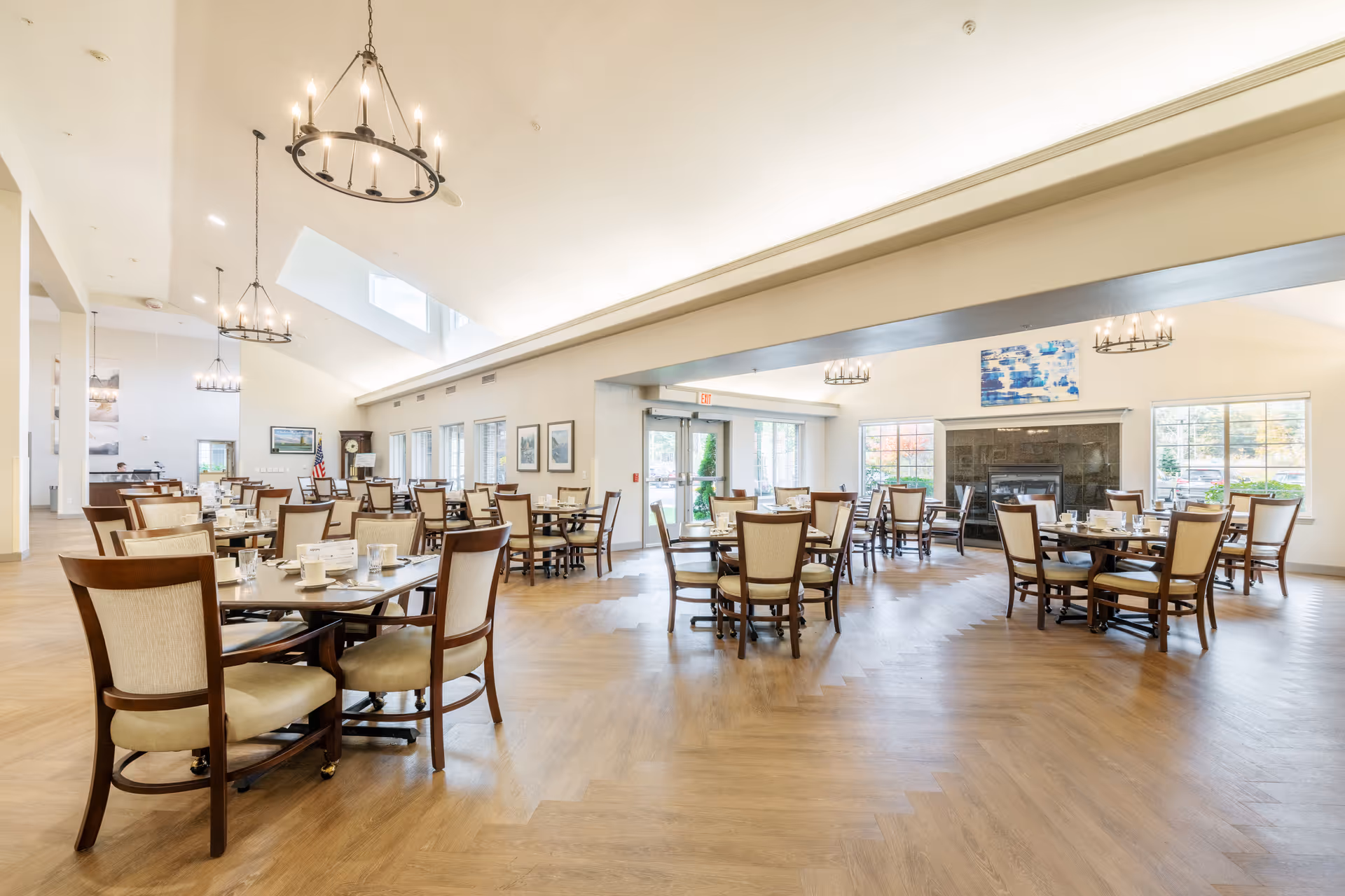 A spacious and well-lit dining room with multiple wooden tables and cushioned chairs arranged neatly. The room features large windows allowing natural light to enter, chandeliers hanging from the ceiling, and a fireplace with a blue and white abstract painting above it. The floor has a light wood pattern, and the overall ambiance is warm and inviting.