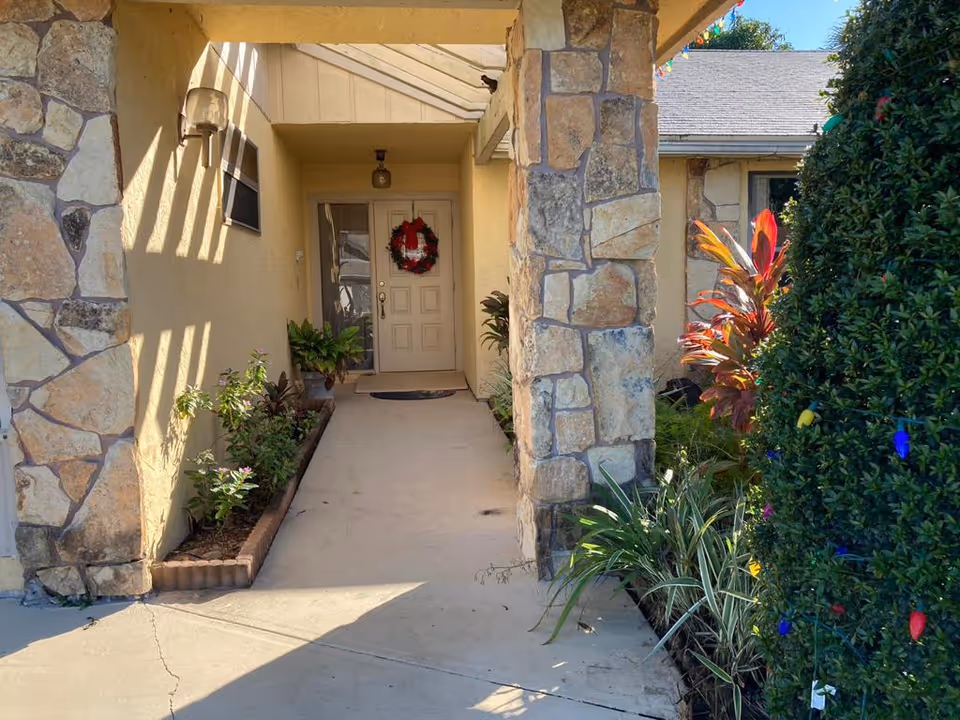Entrance walkway to a building with stone pillars and beige walls, decorated with a red wreath on the front door. There are plants and shrubs along the sides of the walkway, and some colorful lights are visible on the bushes to the right.