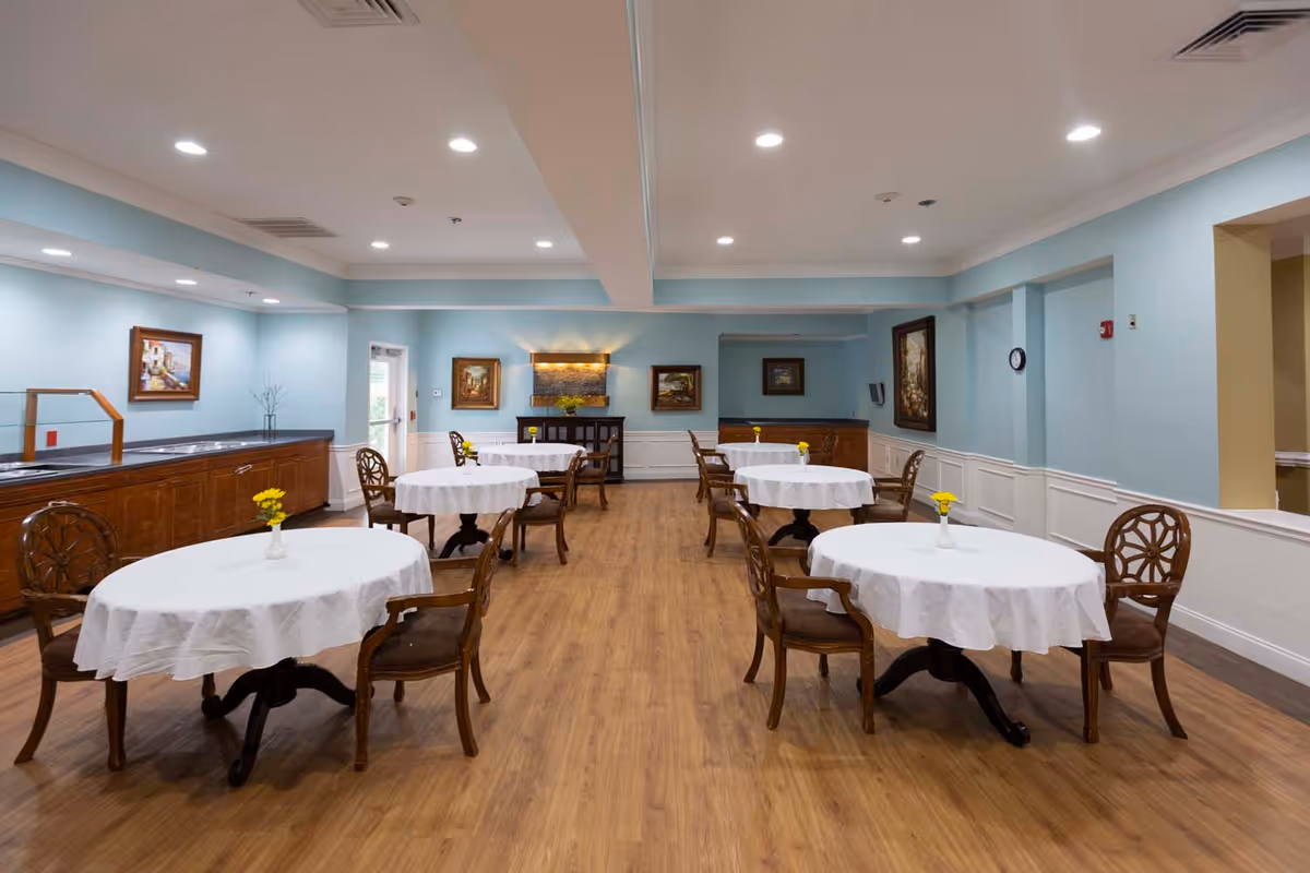 Spacious dining room with round tables covered in white tablecloths, wooden chairs, light blue walls, and wood-look flooring.