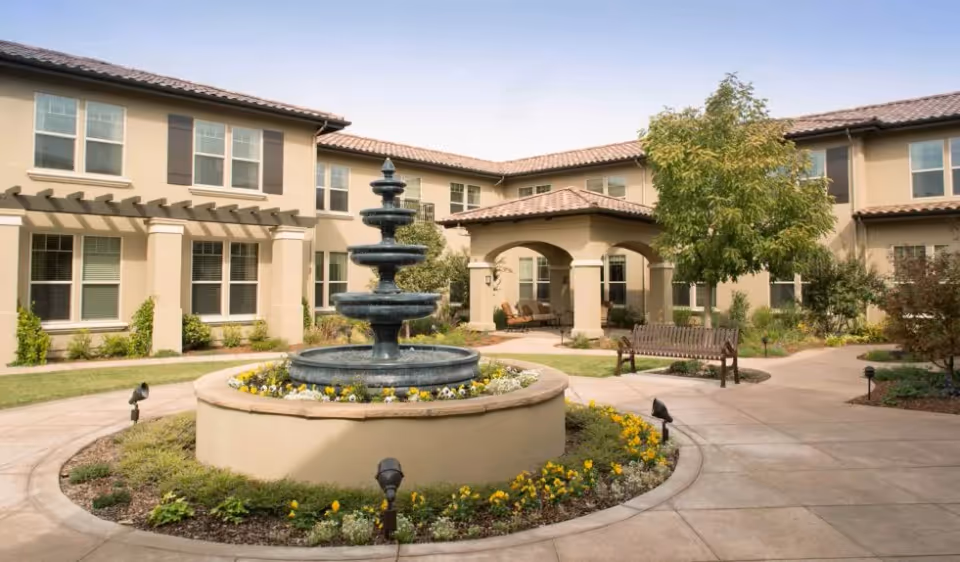 Outdoor courtyard area of a senior living facility with a three-tiered water fountain in the center surrounded by flowers and greenery. The courtyard is paved with walkways and has benches and a covered seating area. The building surrounding the courtyard has beige walls, multiple windows, and a tiled roof.