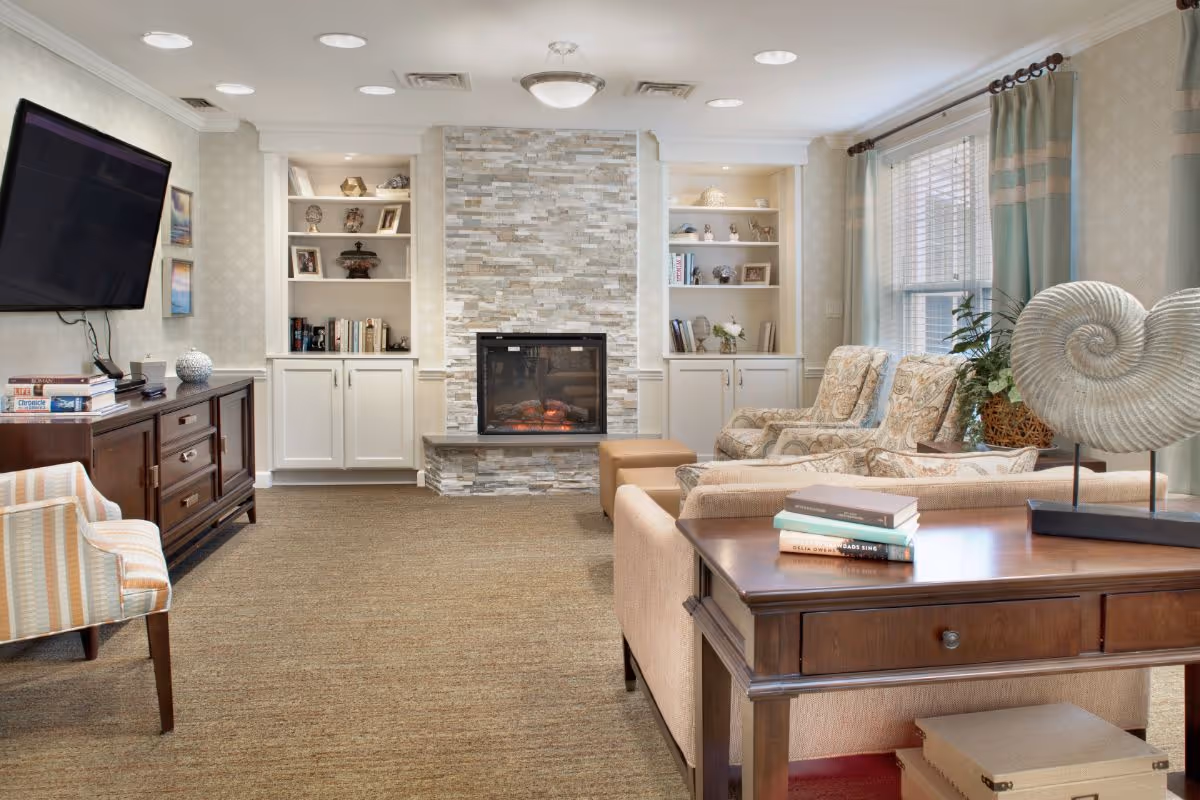 A cozy living room with a stone fireplace in the center, flanked by built-in white shelves and cabinets. There are two patterned armchairs and a beige sofa facing the fireplace. A wooden table behind the sofa holds a decorative shell sculpture and a stack of books. A flat-screen TV is mounted on the left wall above a dark wooden cabinet. The room has large windows with light blue curtains and beige carpeting.