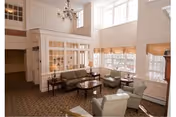A bright and spacious living room area with large windows allowing natural light to fill the space. The room features comfortable seating including a green sofa, two armchairs, and a coffee table in the center. There is a chandelier hanging from the ceiling and a patterned carpet covering the floor. The walls are painted white, and there is a wooden cabinet with glass doors along one side of the room.