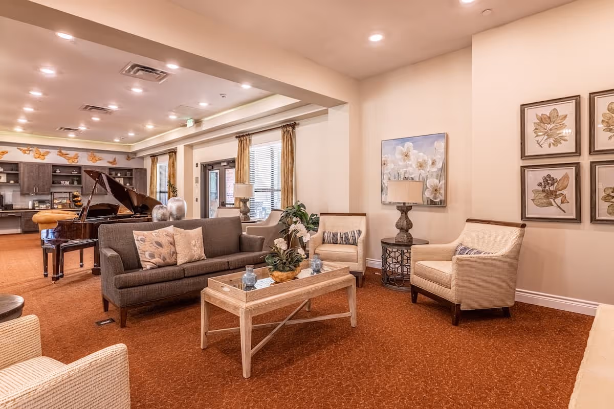 A spacious and well-lit living room area in an assisted living facility featuring a gray sofa with decorative pillows, two beige armchairs, a wooden coffee table with a flower arrangement, and a grand piano in the background. The room has warm lighting, beige walls adorned with floral artwork, and large windows with curtains allowing natural light.