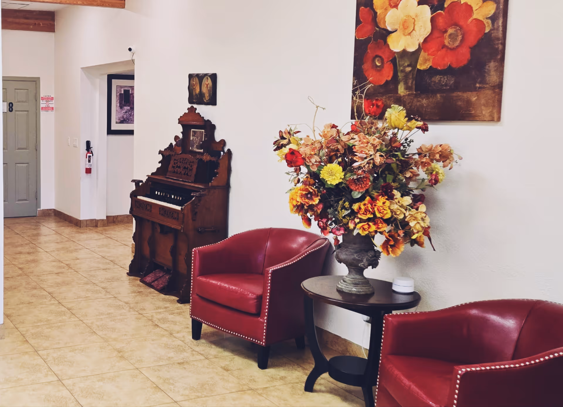 A cozy interior corner of Sunrise Care Homes - Hayden featuring two red leather armchairs with nailhead trim, a small round wooden table with a large floral arrangement in a decorative vase, an antique wooden organ against the wall, and a painting of large colorful flowers hanging above.
