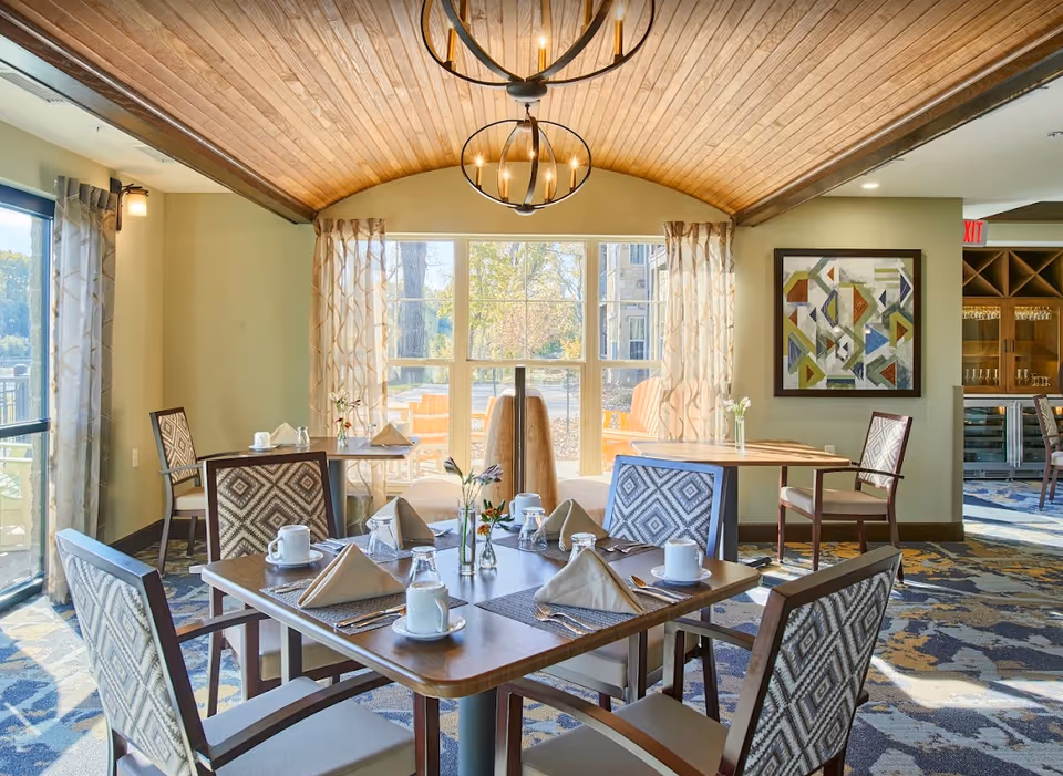 A bright dining room with wooden tables set with cups, glasses, napkins, and silverware. The room features patterned chairs, a curved wooden ceiling with two modern chandeliers, large windows with sheer curtains letting in natural light, and a colorful abstract painting on the wall. Outside, there are orange Adirondack chairs visible through the windows.