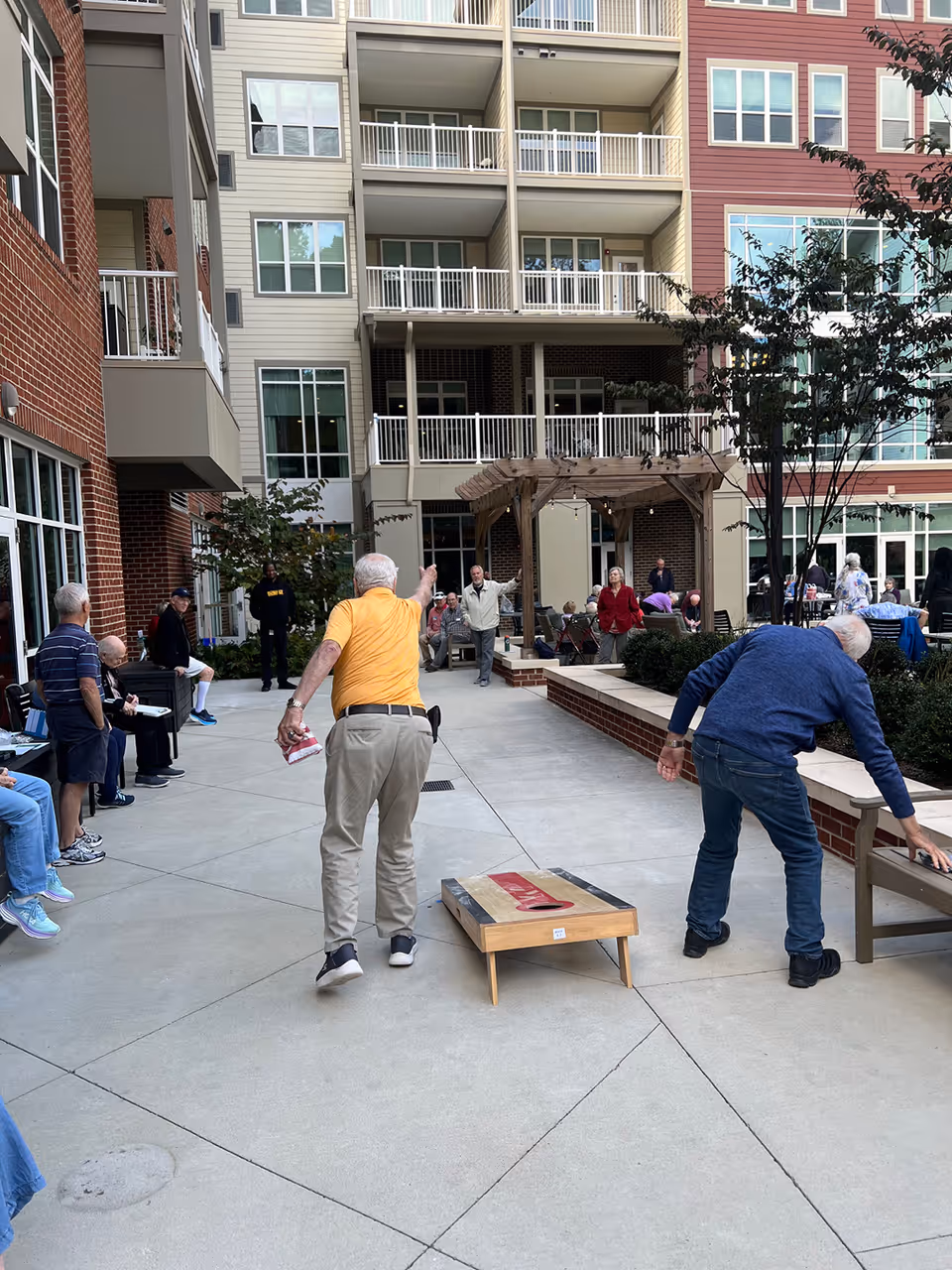 An outdoor courtyard area at a senior living facility with multiple elderly people engaged in activities. Two men are playing a game of cornhole, while others are seated or standing around the courtyard. The background shows a multi-story building with balconies and a wooden pergola.