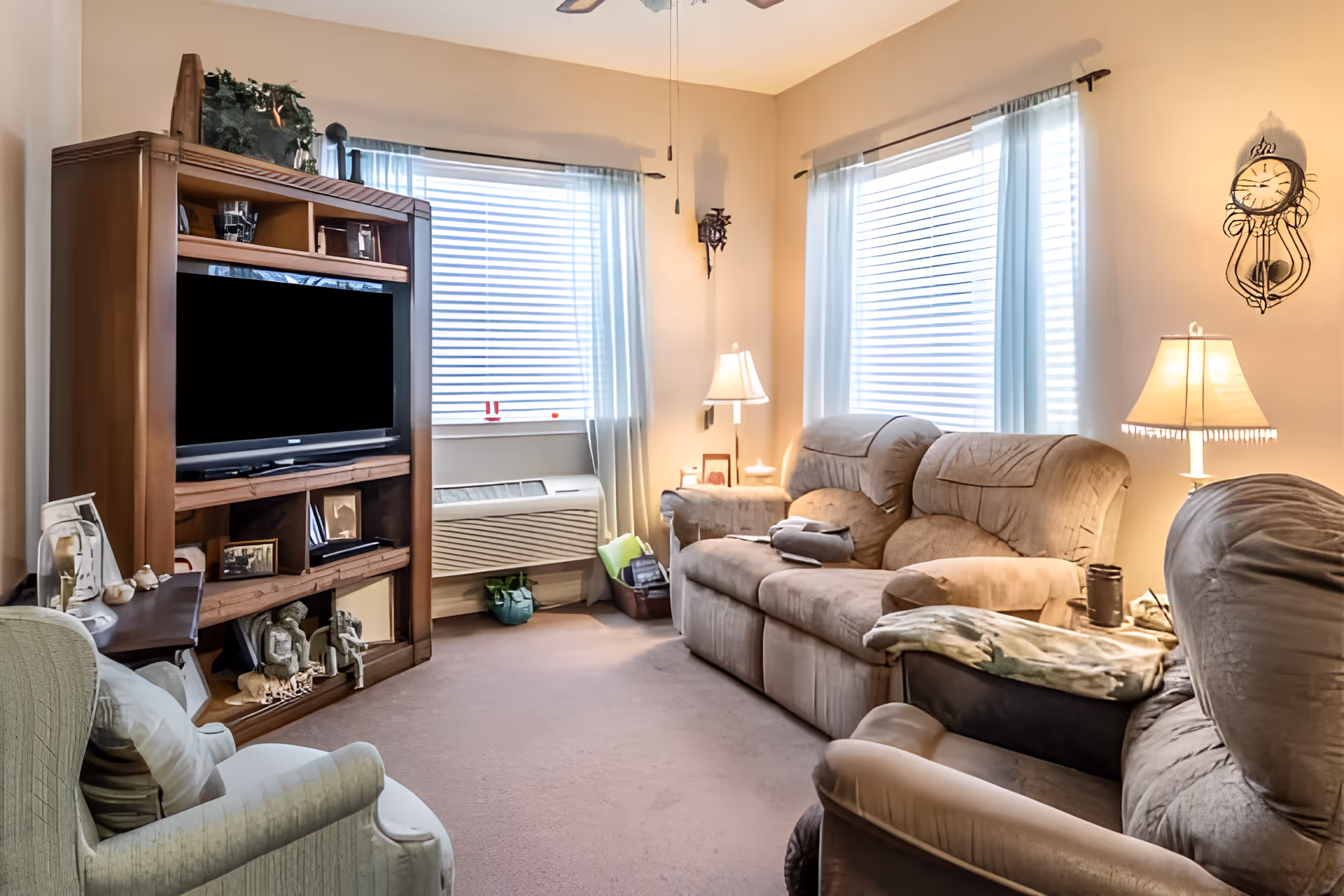 A cozy living room with beige walls and carpet, featuring a large wooden entertainment center with a flat-screen TV, two large windows with white blinds and sheer curtains, a beige reclining sofa, a matching armchair, a white armchair with a pillow, two table lamps, and a decorative wall clock.