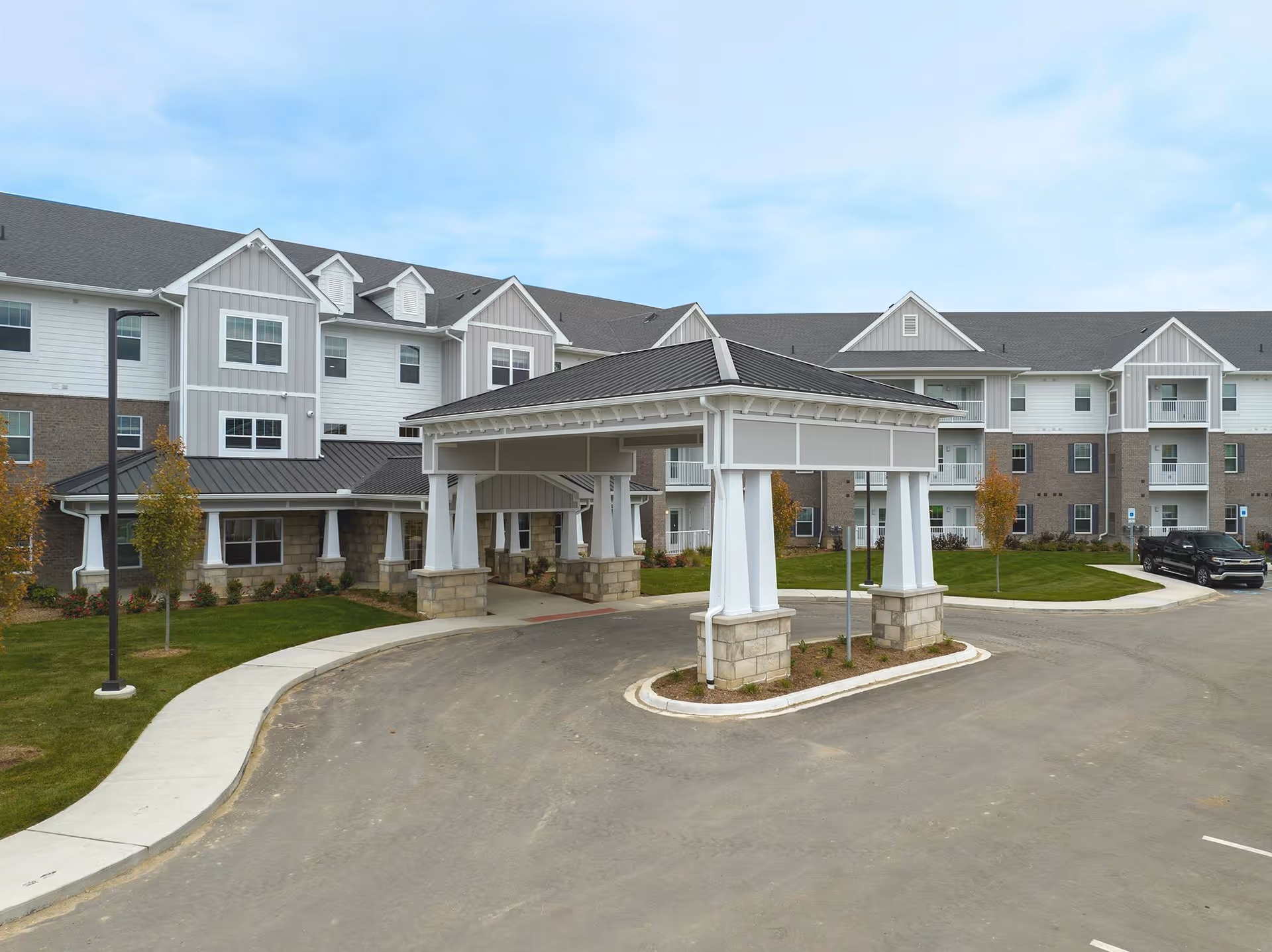 Exterior view of a multi-story senior living facility with a covered entrance driveway, landscaped lawn, and a few small trees under a partly cloudy sky.