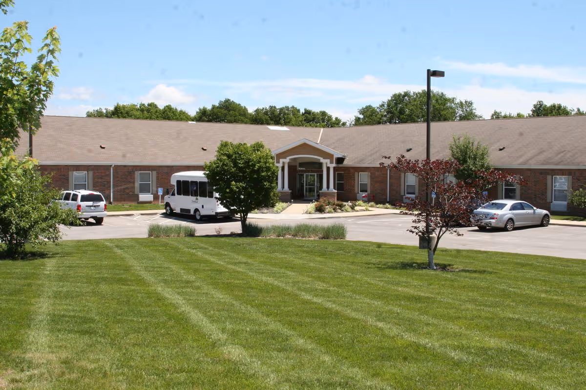Front view of a single-story brick senior living building with a manicured lawn, parking lot, and entrance canopy.