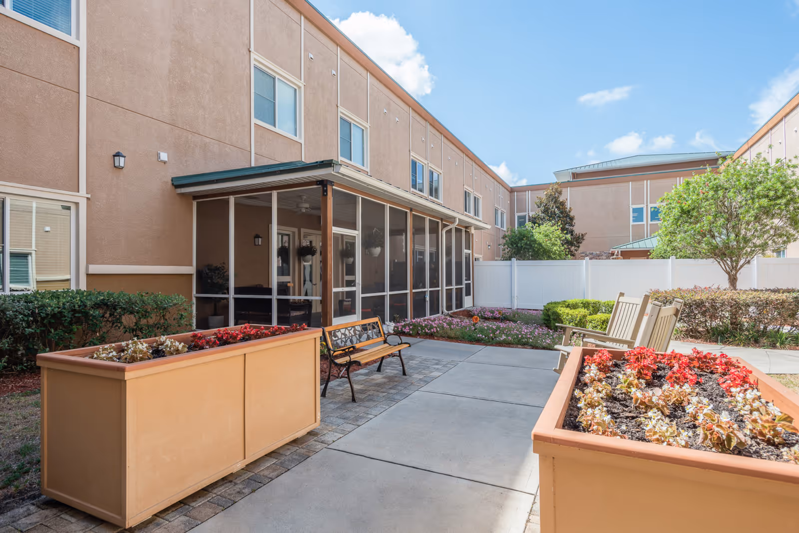 Outdoor courtyard area of a senior living facility with a paved walkway, two large rectangular planters filled with flowers, a wooden bench with metal armrests, and two wooden rocking chairs. The courtyard is surrounded by beige building walls with windows and a screened-in porch. There are bushes, small trees, and a white fence enclosing the area under a partly cloudy blue sky.