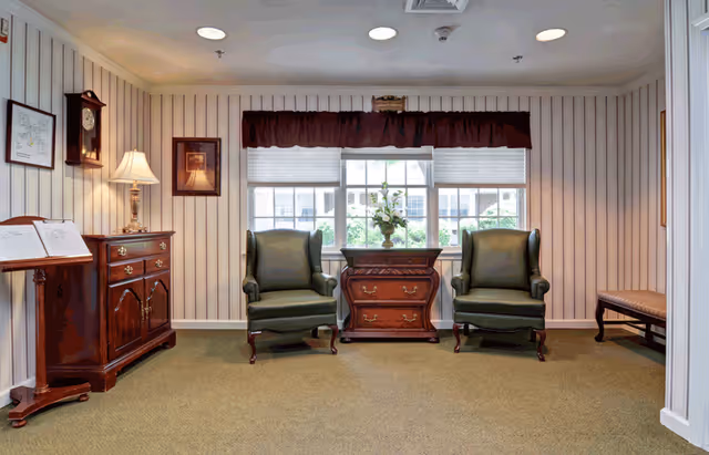 A cozy sitting area with two green upholstered armchairs positioned on either side of a wooden chest with a floral arrangement on top. The room has striped wallpaper, a window with a maroon valance, a wooden sideboard with a lamp and framed pictures on the left, and a wooden bench on the right. The carpet is green and the ceiling has recessed lighting.