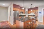 Modern kitchen with wooden cabinets, stainless steel refrigerator, microwave, and stove. A large island with a dark granite countertop has two metal bar stools. Pendant lights hang above the island. The floor is wooden, and there is a red and beige patterned rug near the entrance.