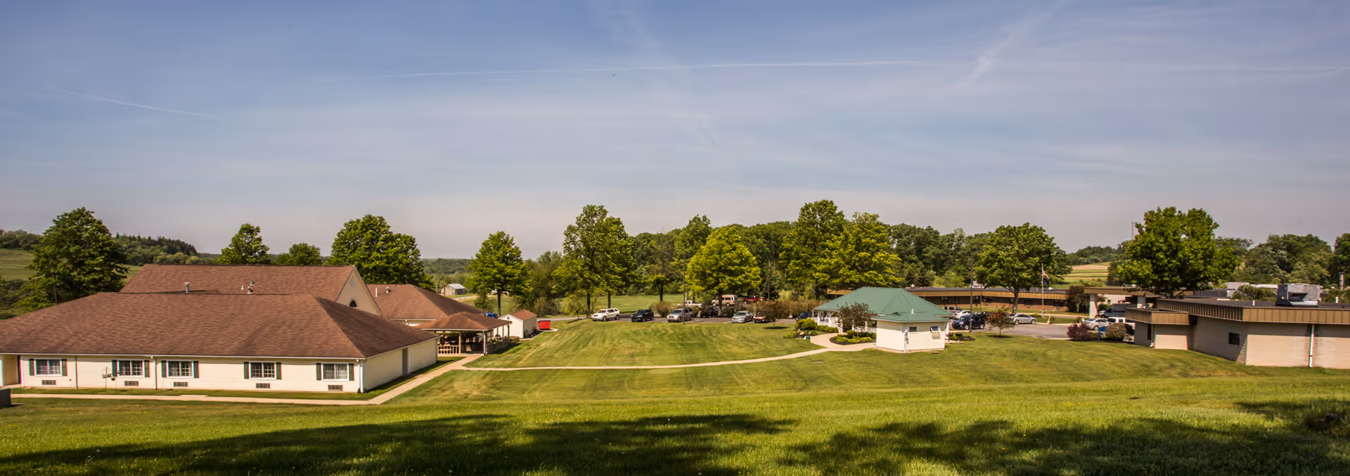 Wide exterior view of Clarview Nursing & Rehab Center showing multiple single-story buildings with brown and green roofs surrounded by a large grassy area and trees under a clear blue sky.