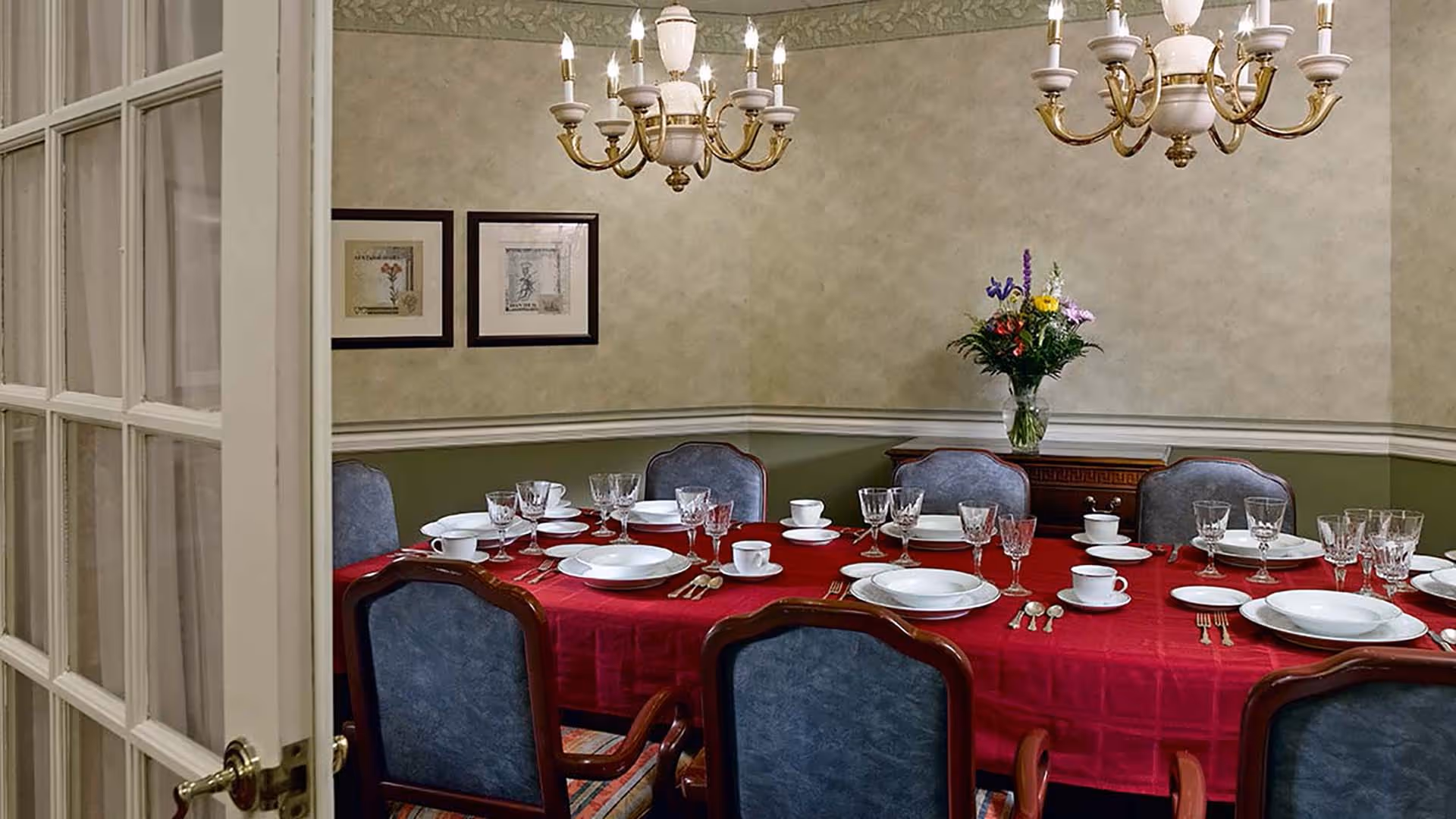 Formal dining room with a red tablecloth, place settings, crystal glassware, upholstered chairs and chandeliers.