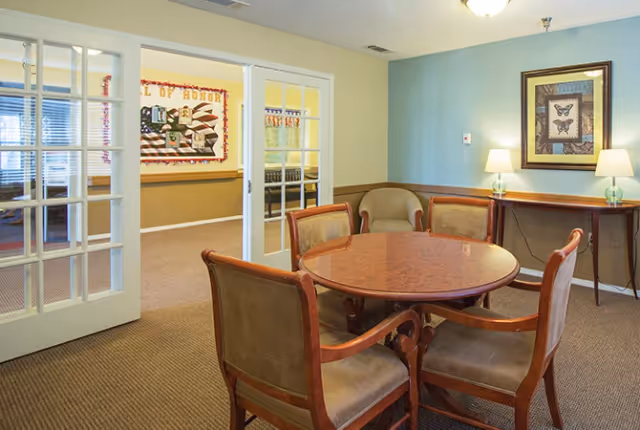 A small common room with a round wooden table and four upholstered chairs, French doors leading to a hallway and a console table with lamps against a blue wall.