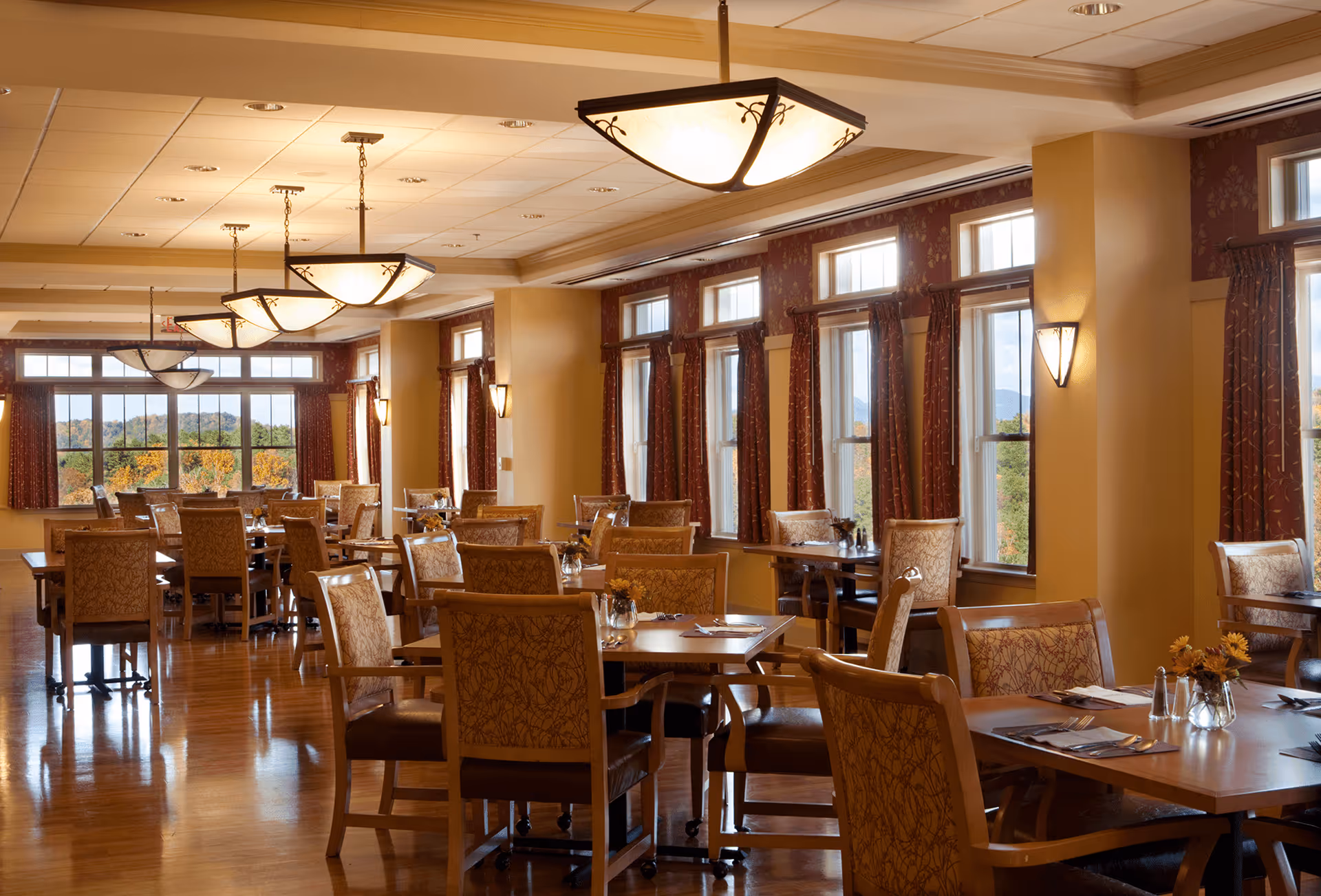 A spacious dining room with multiple wooden tables and cushioned chairs arranged neatly. The room has large windows with red patterned curtains allowing natural light to brighten the space. Elegant ceiling lights and wall sconces provide additional lighting. The floor is polished wood, and small flower arrangements are placed on the tables.