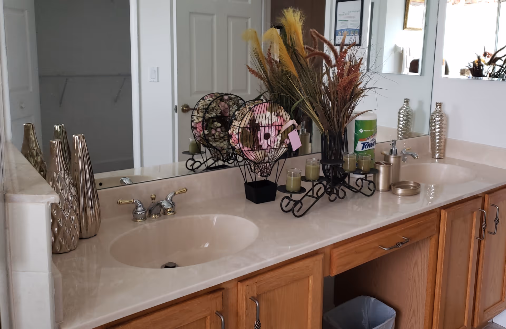 A bathroom vanity with two sinks, a large mirror, and wooden cabinets below. The countertop holds decorative items including metallic vases, a wire basket with decorative balls, a candle holder with multiple candles, a soap dispenser, and a container of paper towels. A door and part of a closet are visible in the background.
