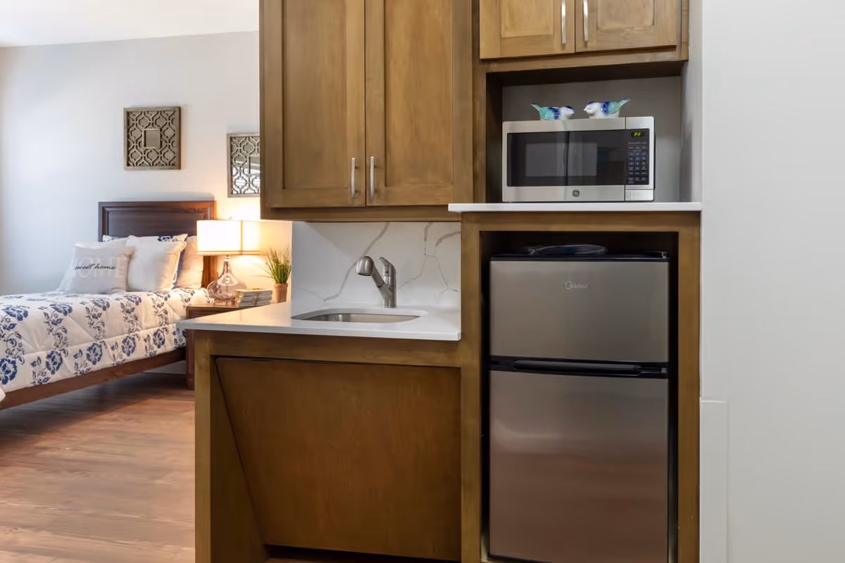 A small kitchenette area with wooden cabinets, a stainless steel mini refrigerator, a microwave, and a sink. In the background, there is a bedroom with a bed featuring a white and blue floral comforter, a nightstand with a lamp, and decorative wall art.