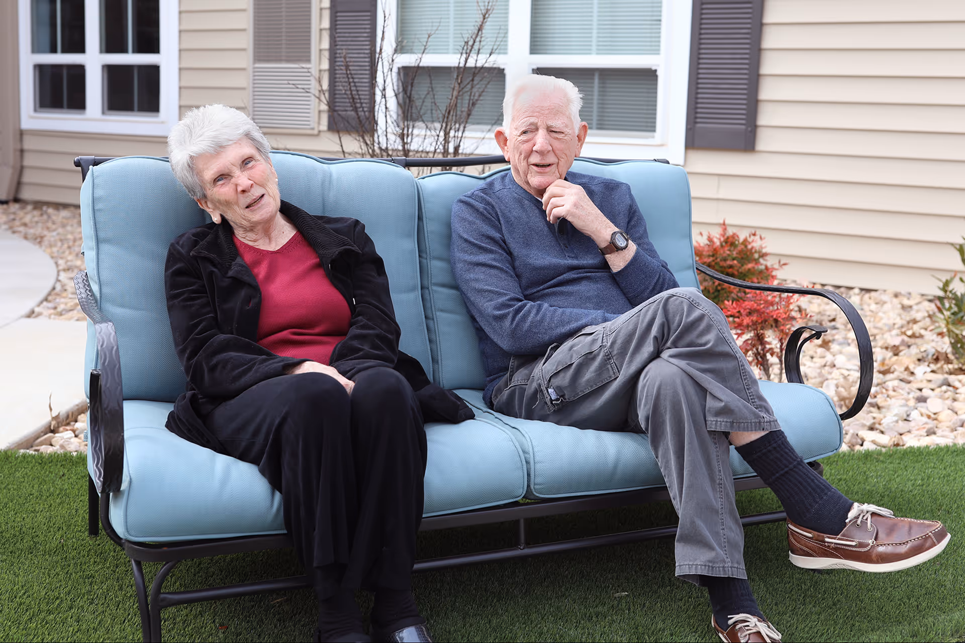 An elderly man and woman sitting on a blue cushioned outdoor bench in a garden area with artificial grass, rocks, and plants. The man is wearing a blue sweater, gray pants, and brown shoes, while the woman is wearing a black jacket, red shirt, and black pants. They are seated in front of a beige building with windows and shutters.