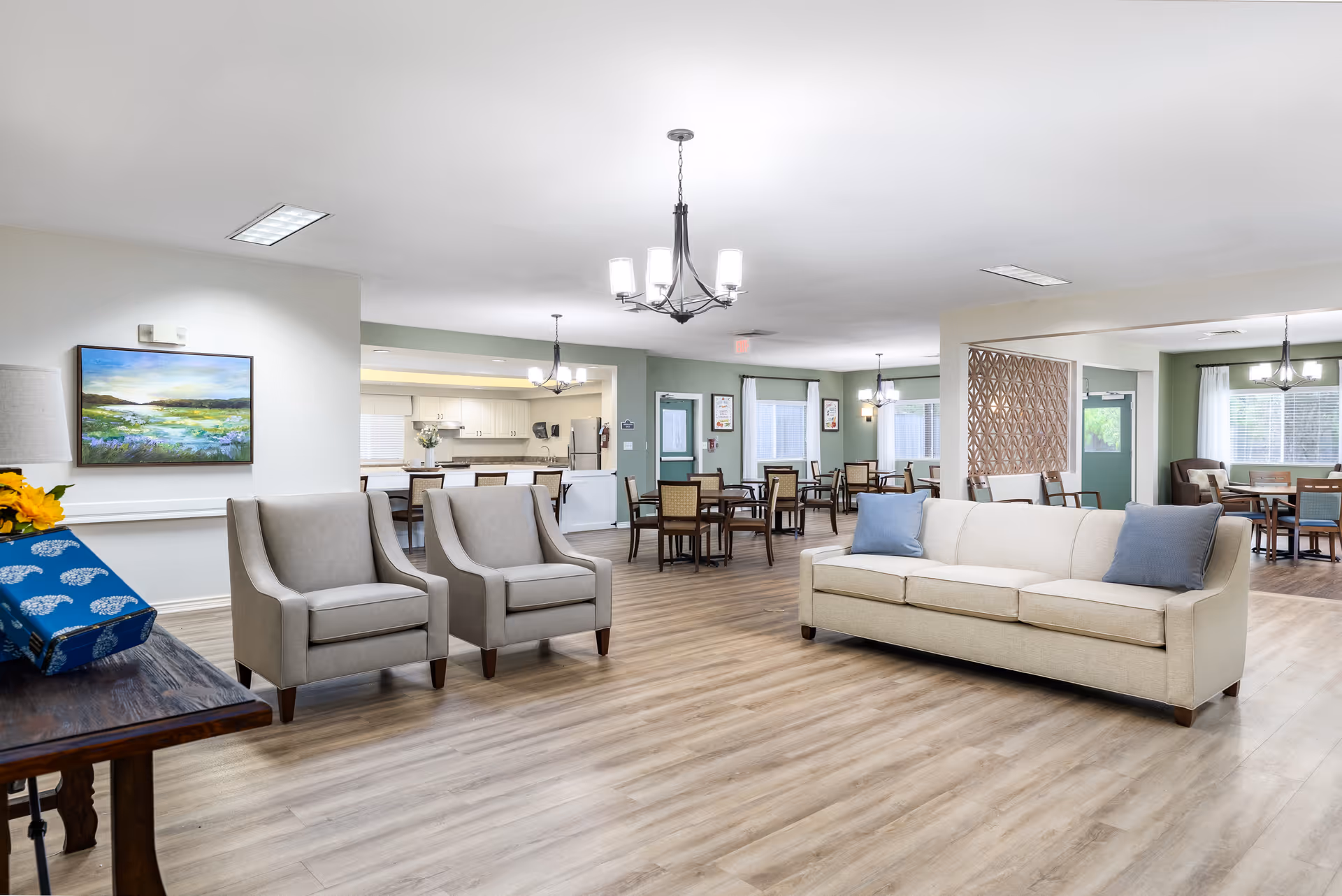 A spacious and well-lit senior living common area with light wood flooring, beige sofa with blue pillows, two gray armchairs, multiple dining tables with chairs, and a kitchen area in the background. The walls are painted in soft green and white tones, and there are several ceiling light fixtures and large windows with white curtains.