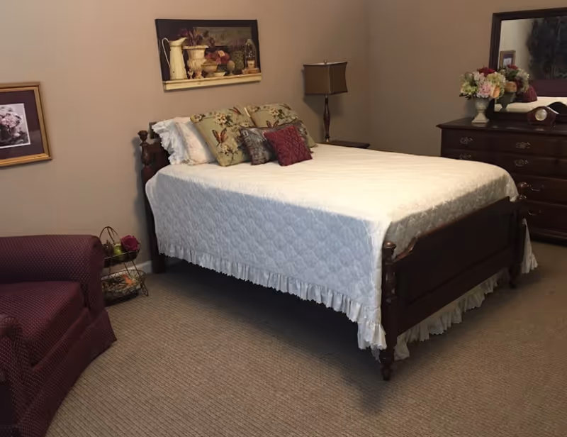 A cozy bedroom with a wooden bed frame covered in a white quilt and decorative pillows. There is a wooden dresser with a mirror and a vase of flowers on top. A floor lamp stands beside the bed, and a maroon upholstered chair is positioned in the corner. The walls are decorated with framed artwork.