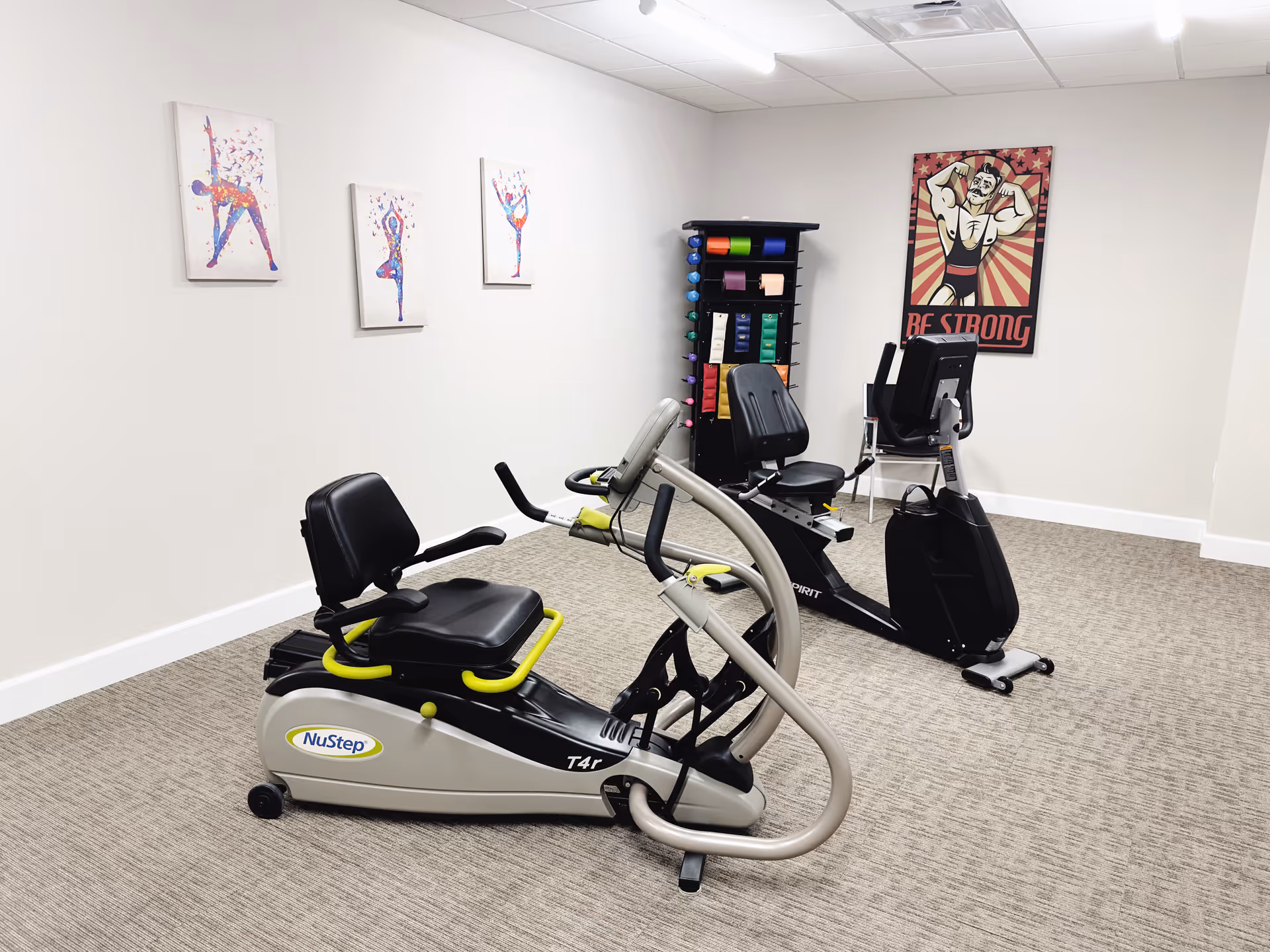 A small exercise room with two recumbent exercise bikes on a carpeted floor. The walls are white and decorated with three colorful paintings of people in yoga poses and a poster of a muscular cartoon man with the text 'BE STRONG'. There is also a rack holding various exercise bands and equipment.