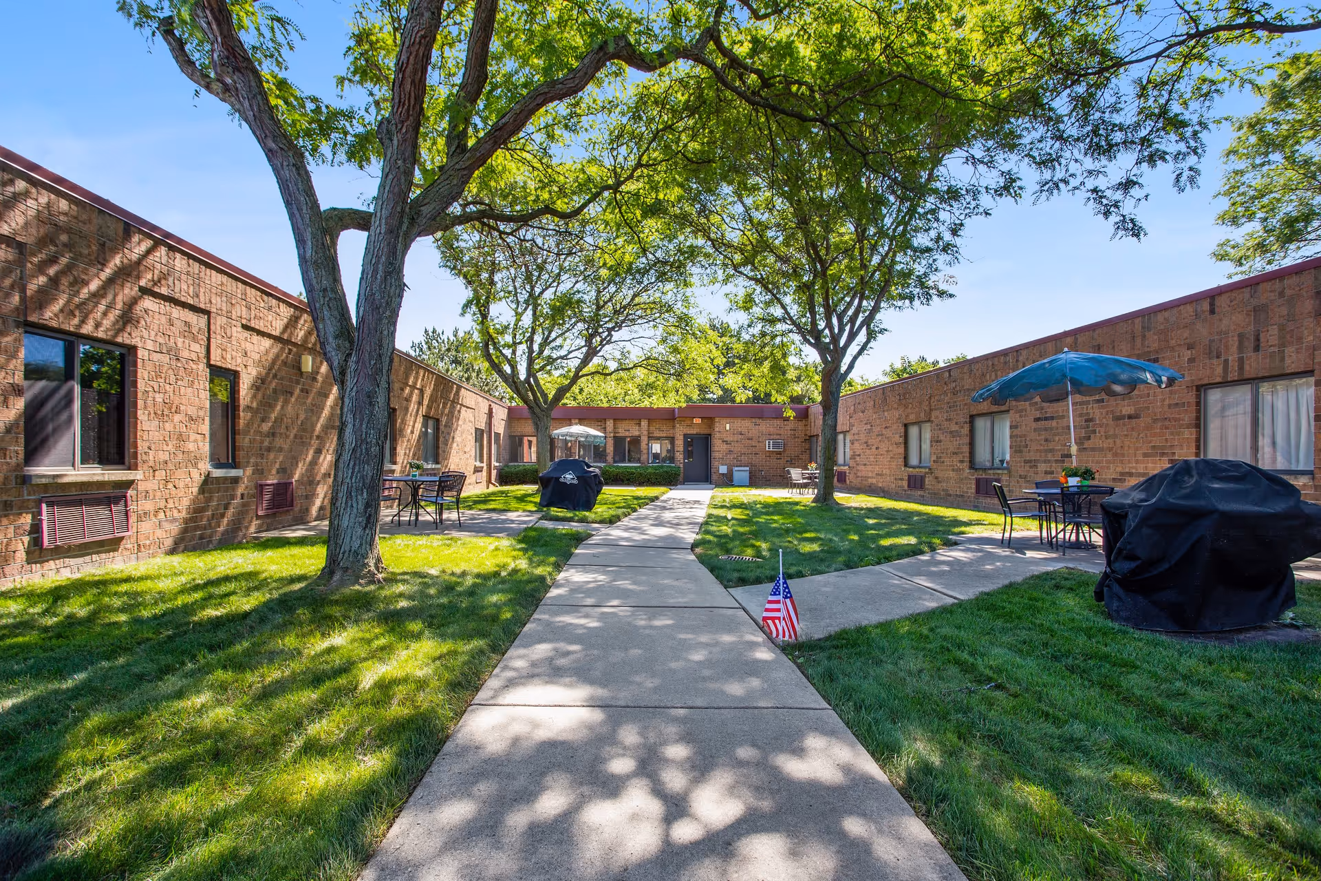 Outdoor courtyard area of a senior living facility with a concrete walkway, green grass, trees providing shade, patio tables with umbrellas, and barbecue grills. The surrounding building is a single-story brick structure with several windows.