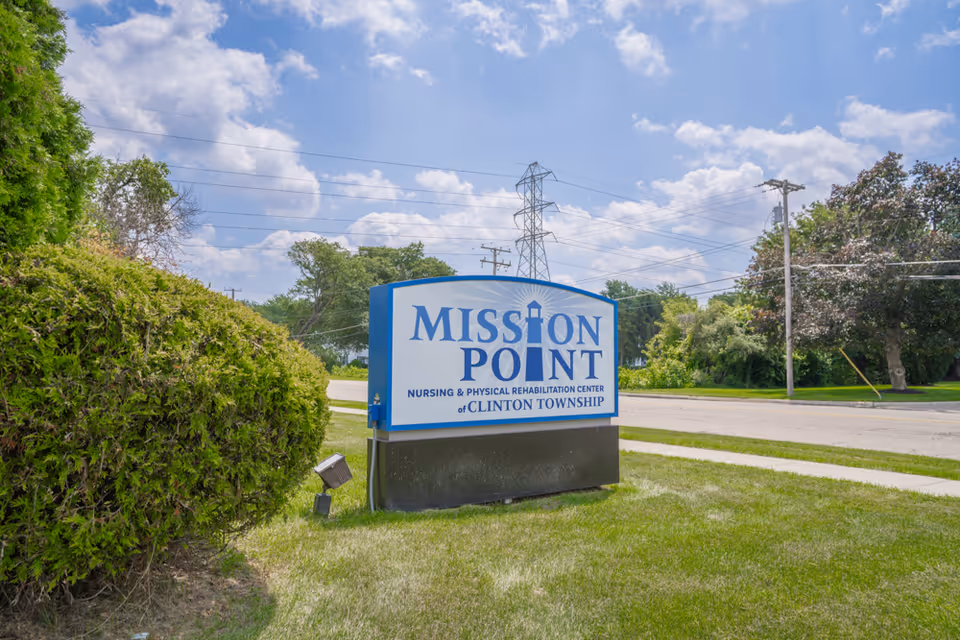 Outdoor view of a sign for Mission Point Nursing & Physical Rehabilitation Center of Clinton Township, situated on a grassy area with bushes and trees nearby under a partly cloudy sky.
