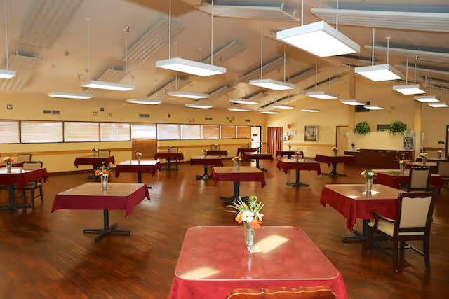 A spacious dining room with multiple tables covered in red tablecloths, each adorned with a small vase of flowers. The room has wooden flooring, large windows along one wall, and modern rectangular ceiling lights evenly spaced across the ceiling.