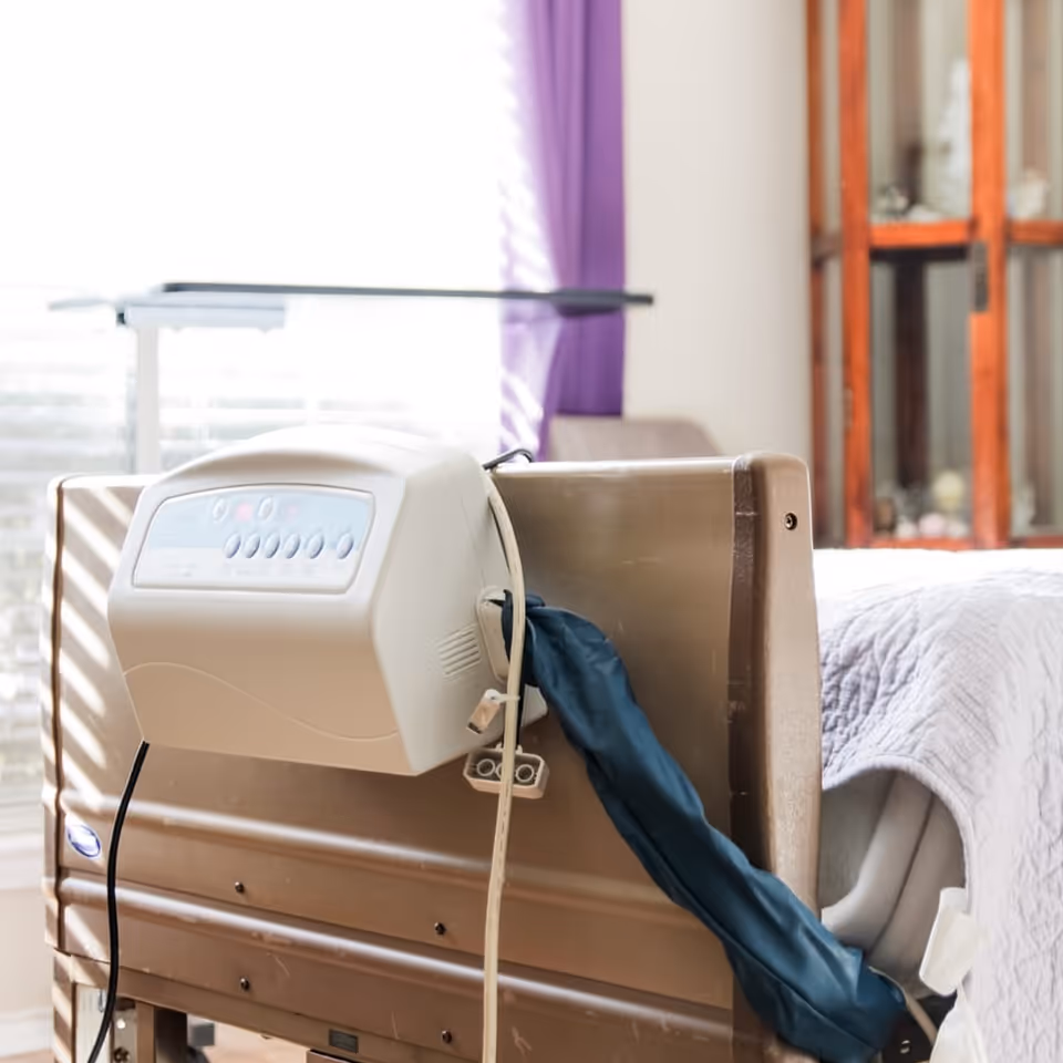 A medical compression device clipped to a bed headboard in a sunlit bedroom with purple curtains and a wooden cabinet.