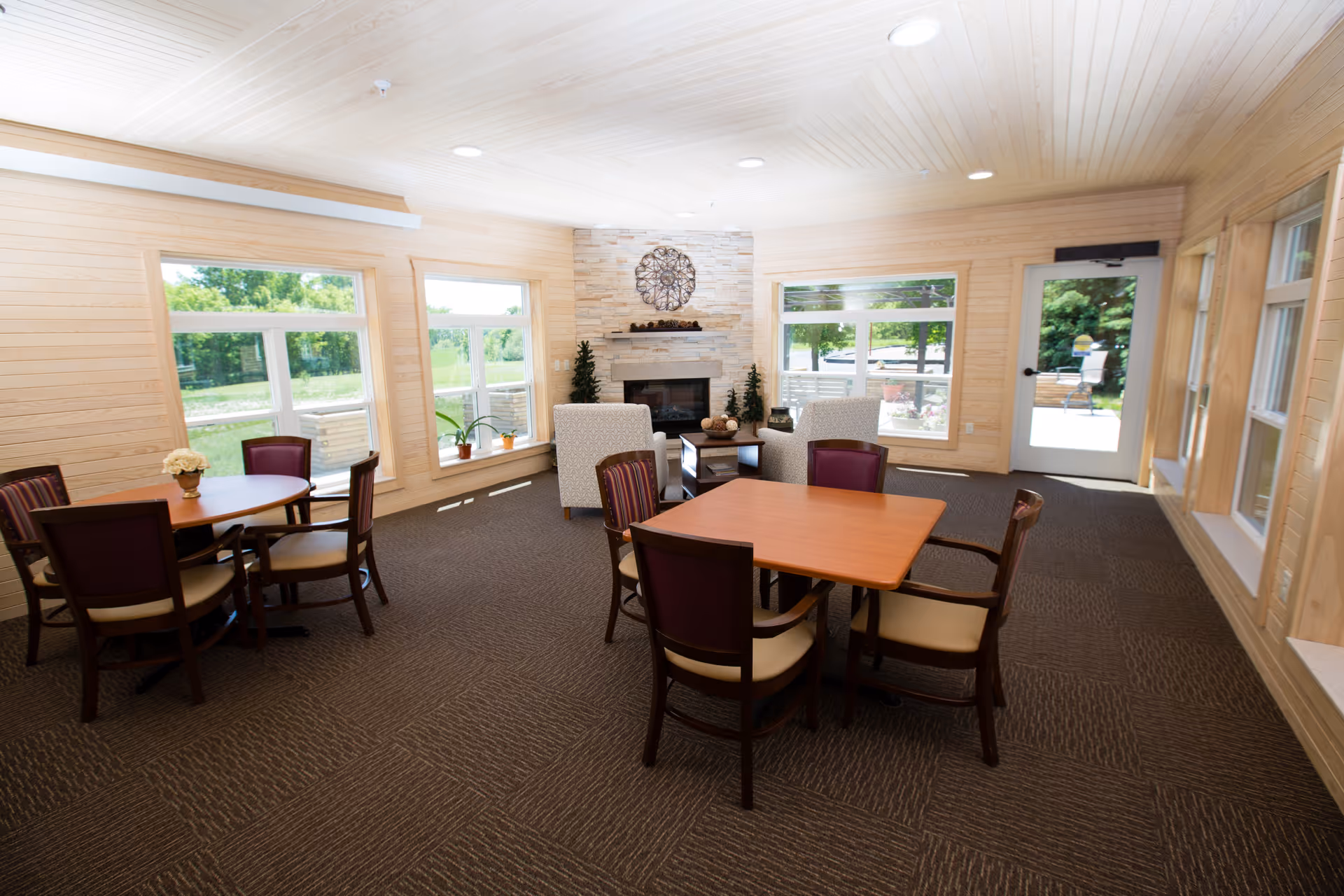 A bright and spacious common area with large windows letting in natural light. The room features two wooden tables with chairs around them, two upholstered armchairs near a stone fireplace, and a door leading outside. The walls and ceiling are covered with light wood paneling, and the floor is carpeted in a dark pattern.