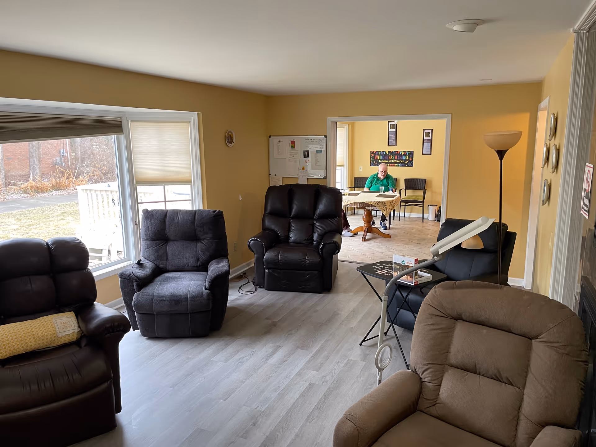 A cozy senior living facility common area with several recliner chairs arranged around the room. There is a large window on the left letting in natural light. In the background, a man is sitting at a table in an adjoining room with yellow walls and a motivational poster on the wall.