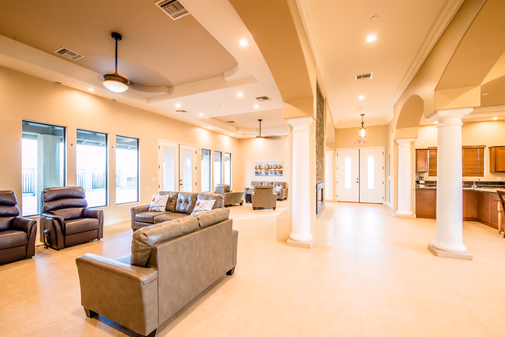 A spacious and well-lit living area in an assisted living facility featuring multiple leather sofas and recliners arranged for socializing. The room has large windows allowing natural light, ceiling fans, recessed lighting, and beige tiled floors. White columns and arches separate the living area from an adjacent kitchen with wooden cabinets and granite countertops.