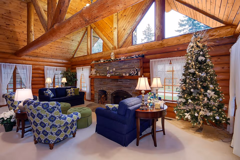 A decorated log-cabin living room with a stone fireplace, Christmas tree, and upholstered sofas and armchairs under a vaulted wood ceiling.
