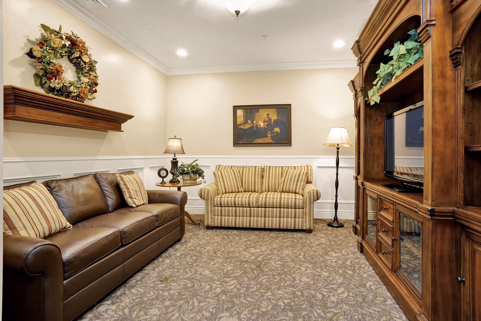 A cozy living room with a brown leather sofa and a striped loveseat. There is a wooden entertainment center with a TV on the right side, a floor lamp, and a table lamp on a small round table between the sofas. A decorative wreath hangs on the wall above the leather sofa, and a framed painting is centered on the wall above the loveseat. The room has beige walls with white wainscoting and a patterned carpet.