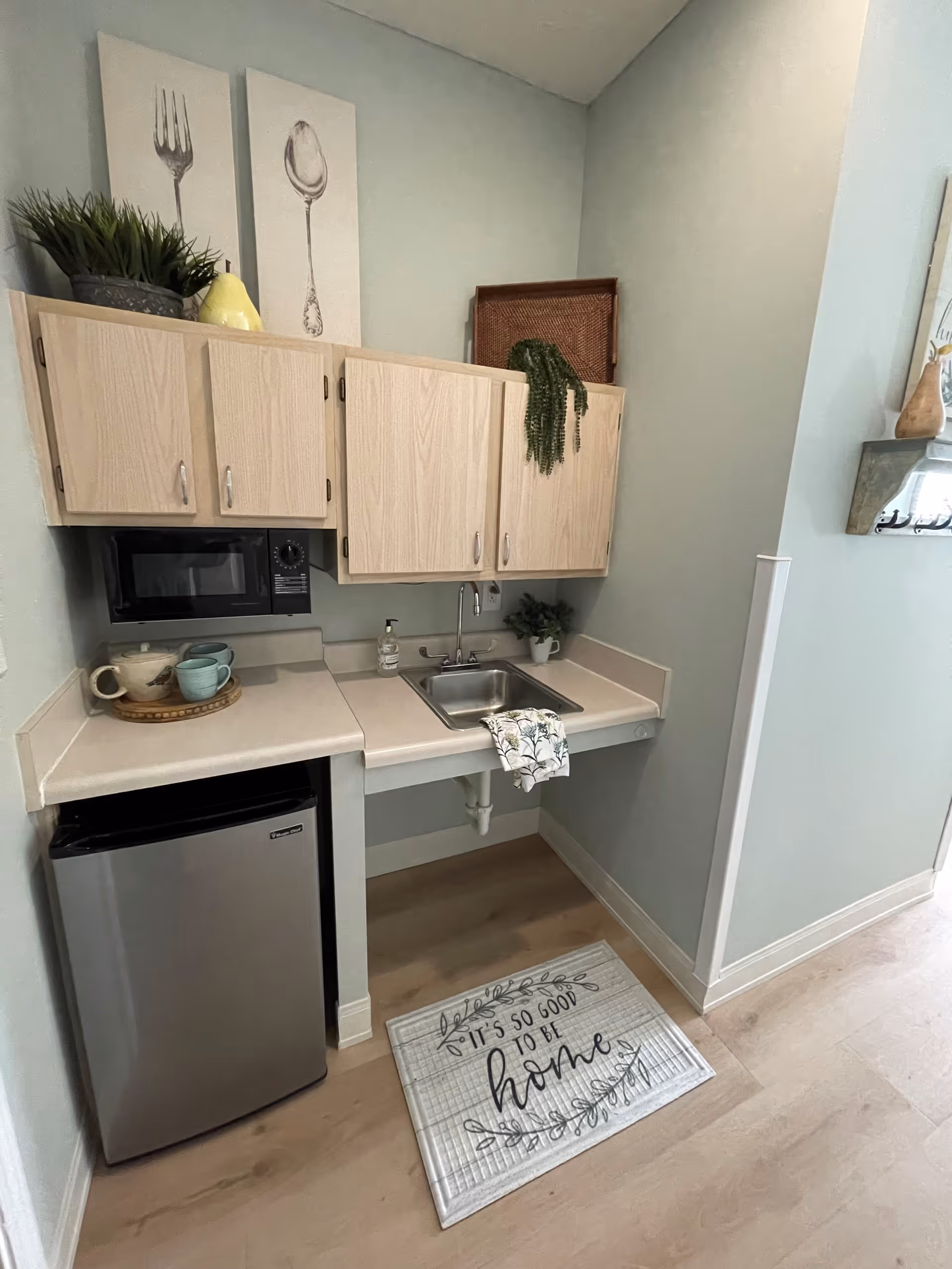 A small kitchenette area with light wood cabinets, a stainless steel mini refrigerator, a microwave, and a sink. There are decorative items on top of the cabinets including a plant and artwork featuring a fork and spoon. A dish towel hangs over the sink and a floor mat in front of the kitchenette reads 'It's so good to be home.'